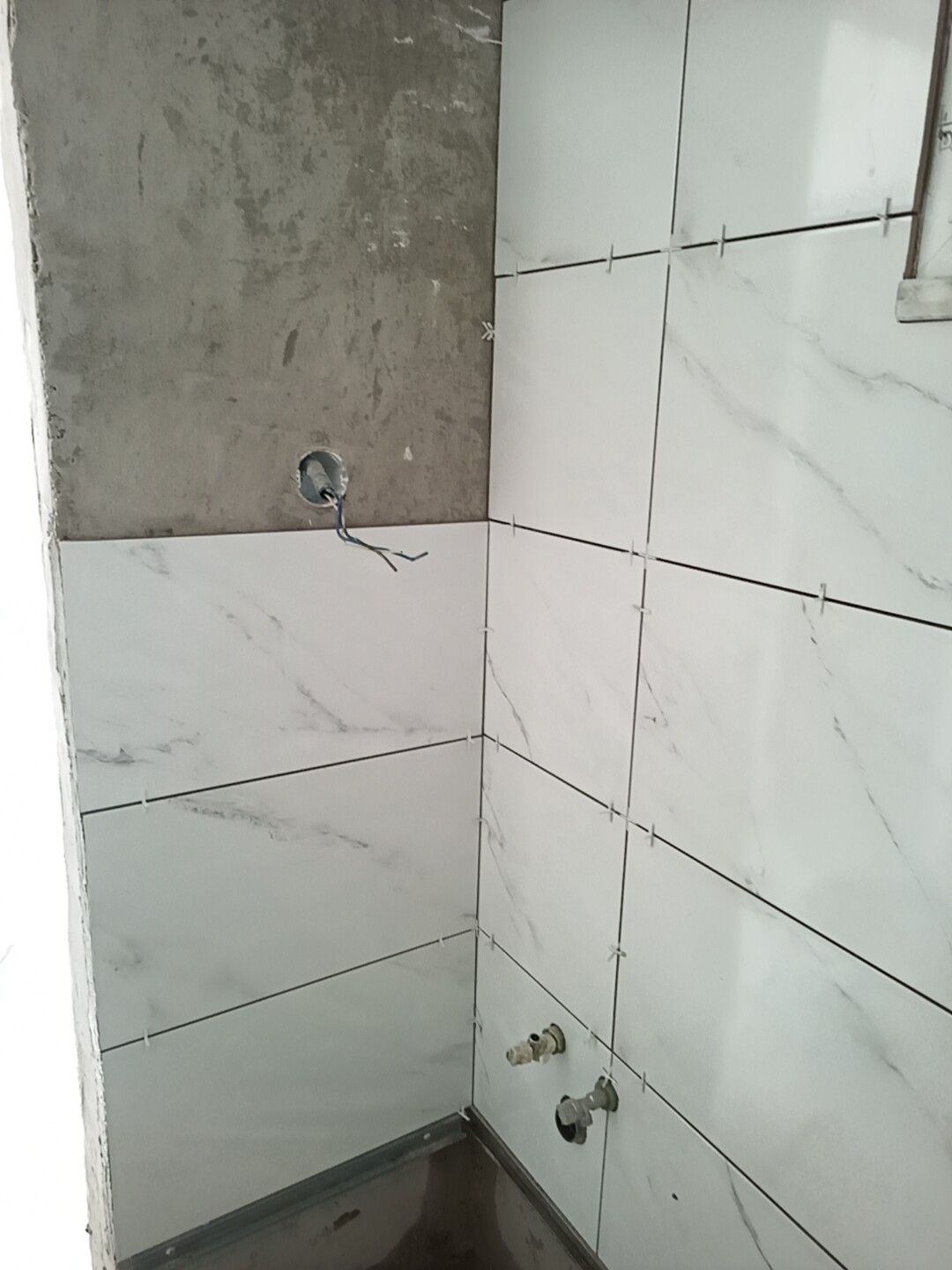 Canto de um banheiro em construção, com azulejos brancos imitando mármore instalados em duas paredes e parede de concreto inacabado.