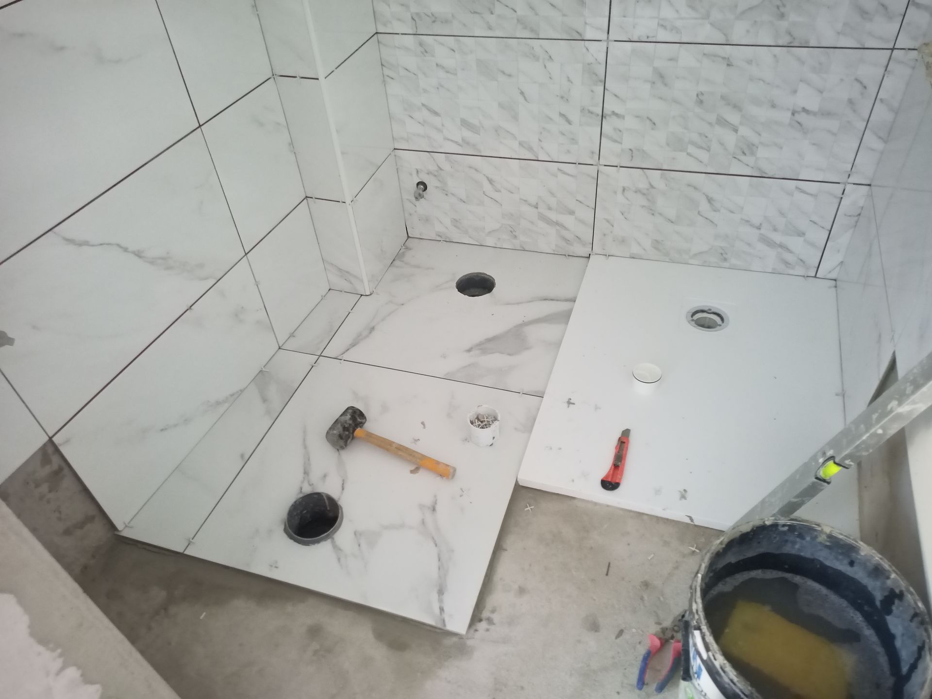 Banheiro em construção com instalação de azulejos brancos imitando mármore. Martelo, balde e ferramentas estão presentes.