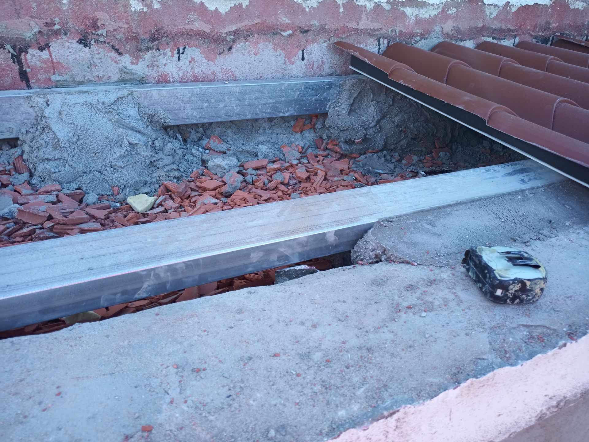 Detalhe de um canteiro de obras: canaleta metálica, concreto, telhas e entulho de tijolos. Uma ferramenta de medição é visível.