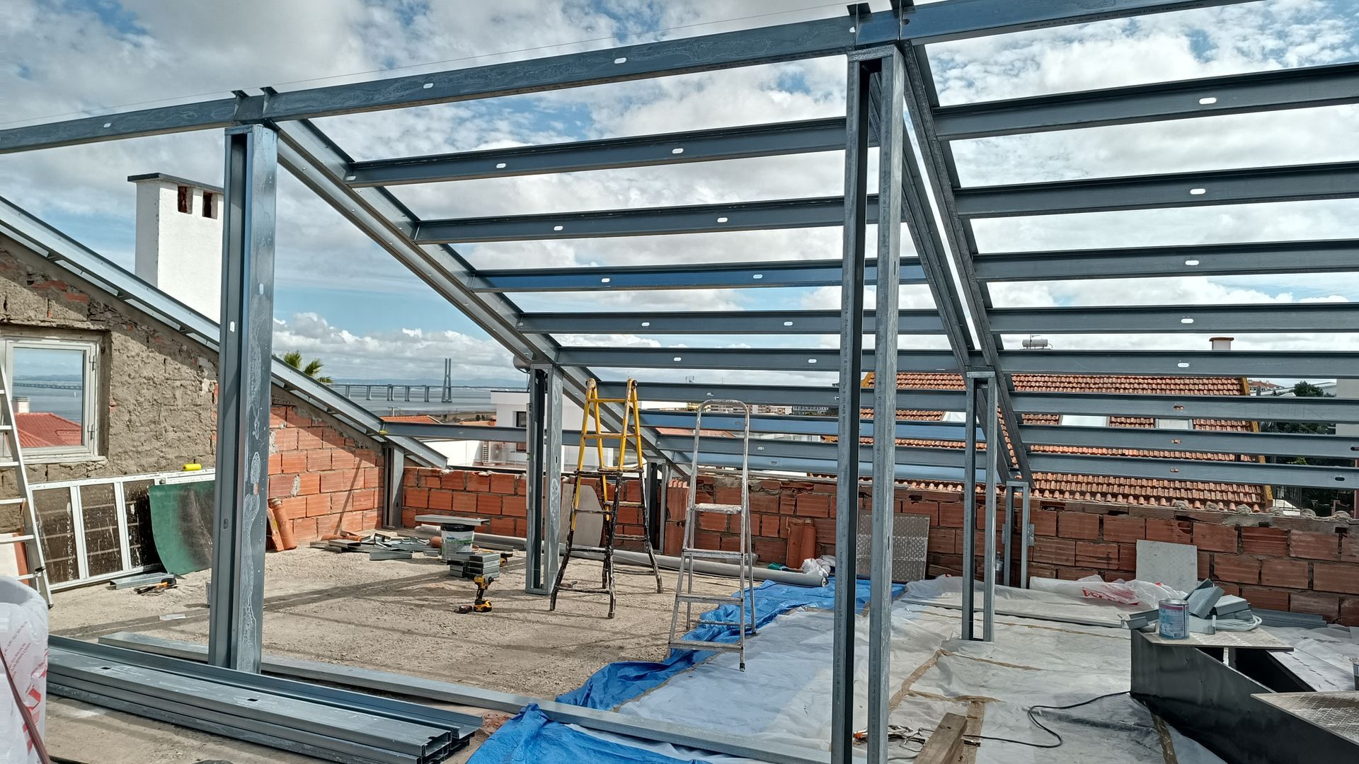 Estrutura de aço que forma uma área externa coberta no terraço de um edifício; tijolos, lona azul, paisagem urbana ao fundo.