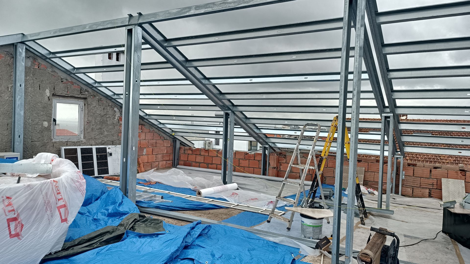 Canteiro de obras: telhado com estrutura metálica, paredes de tijolo, lona azul, escada, céu nublado.