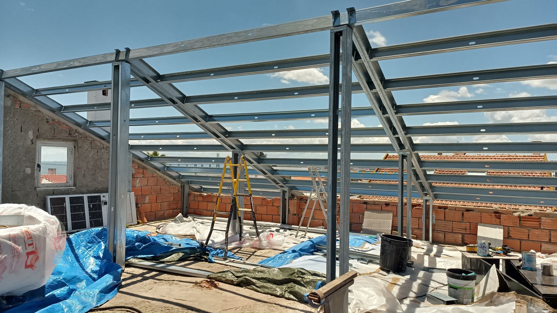 Construção de um telhado com estrutura metálica sobre um edifício de tijolos, com ferramentas e materiais espalhados ao redor; céu azul.