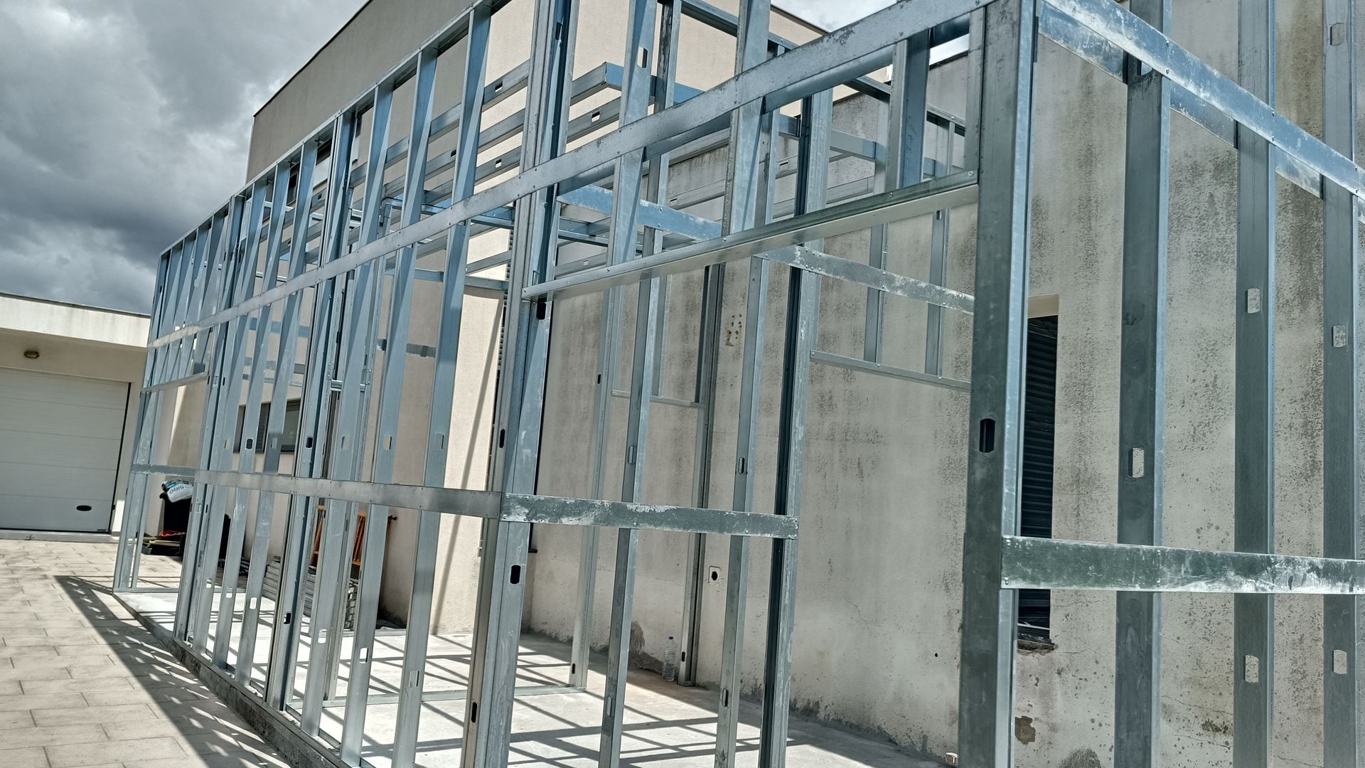 Estrutura metálica de um edifício em construção, contra uma parede branca e céu nublado.
