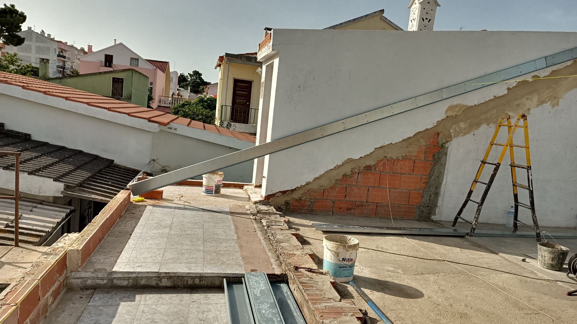 Canteiro de obras com tijolos aparentes e vigas metálicas em um telhado, dia ensolarado.