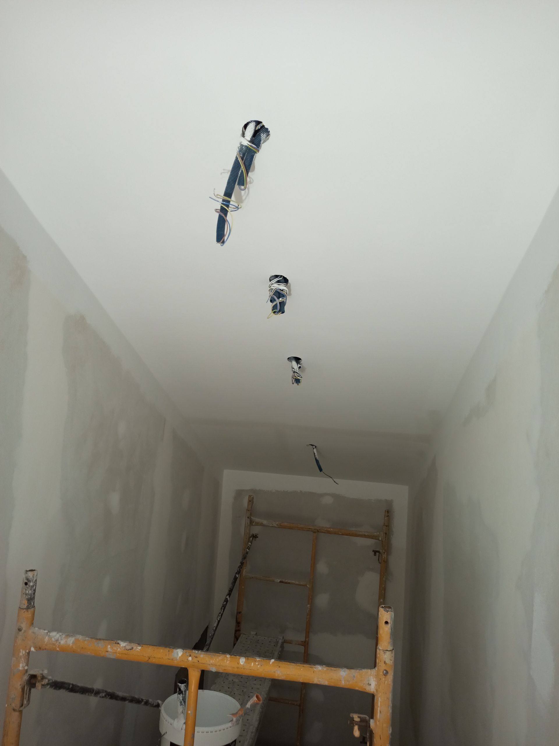 Corredor com paredes e teto inacabados; fios elétricos expostos; andaimes de madeira.