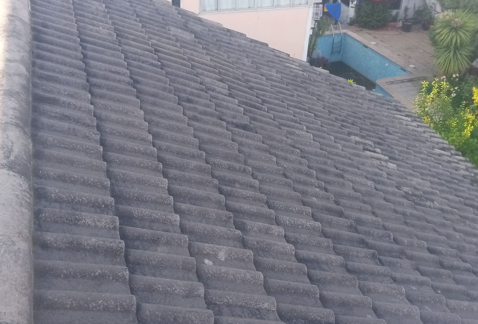 Close-up de um telhado de telhas cinza-escuras desgastado pelo tempo, com uma piscina visível ao fundo.