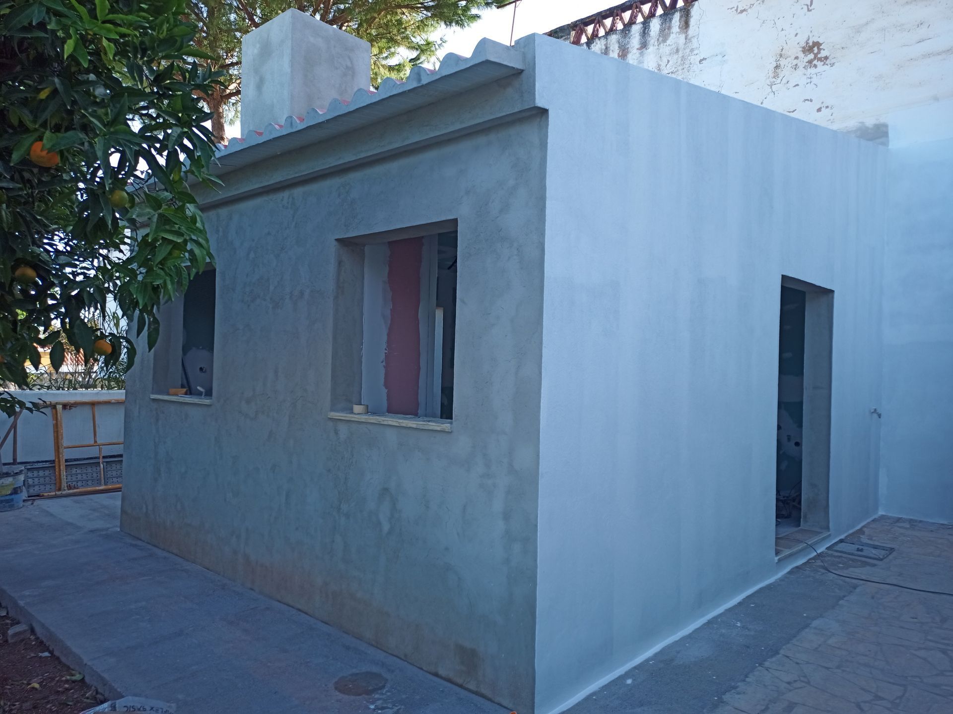 Vista exterior de um pequeno edifício com paredes cinzentas, janela e porta, com uma árvore ao fundo.