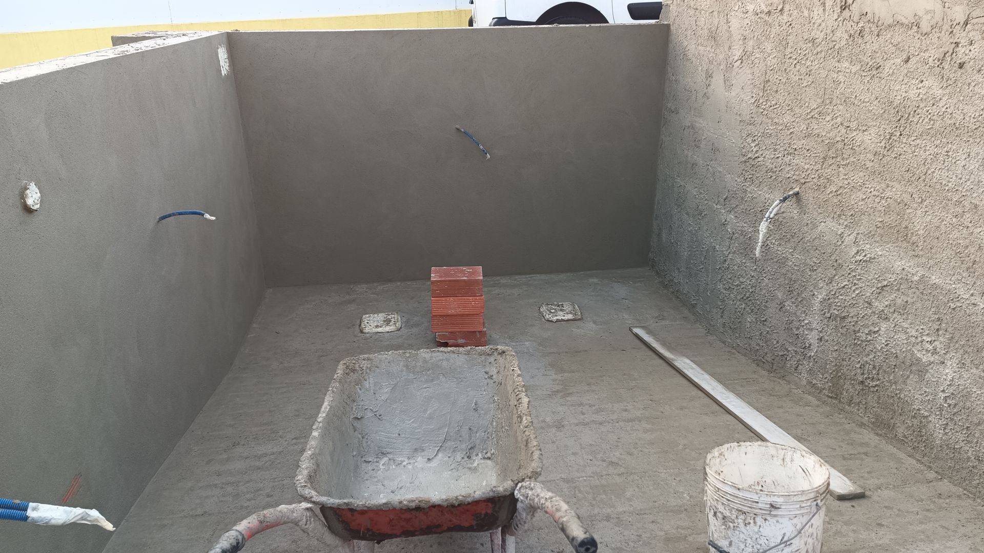 Sala de concreto vazia em construção, com um carrinho de mão, tijolos e ferramentas presentes.