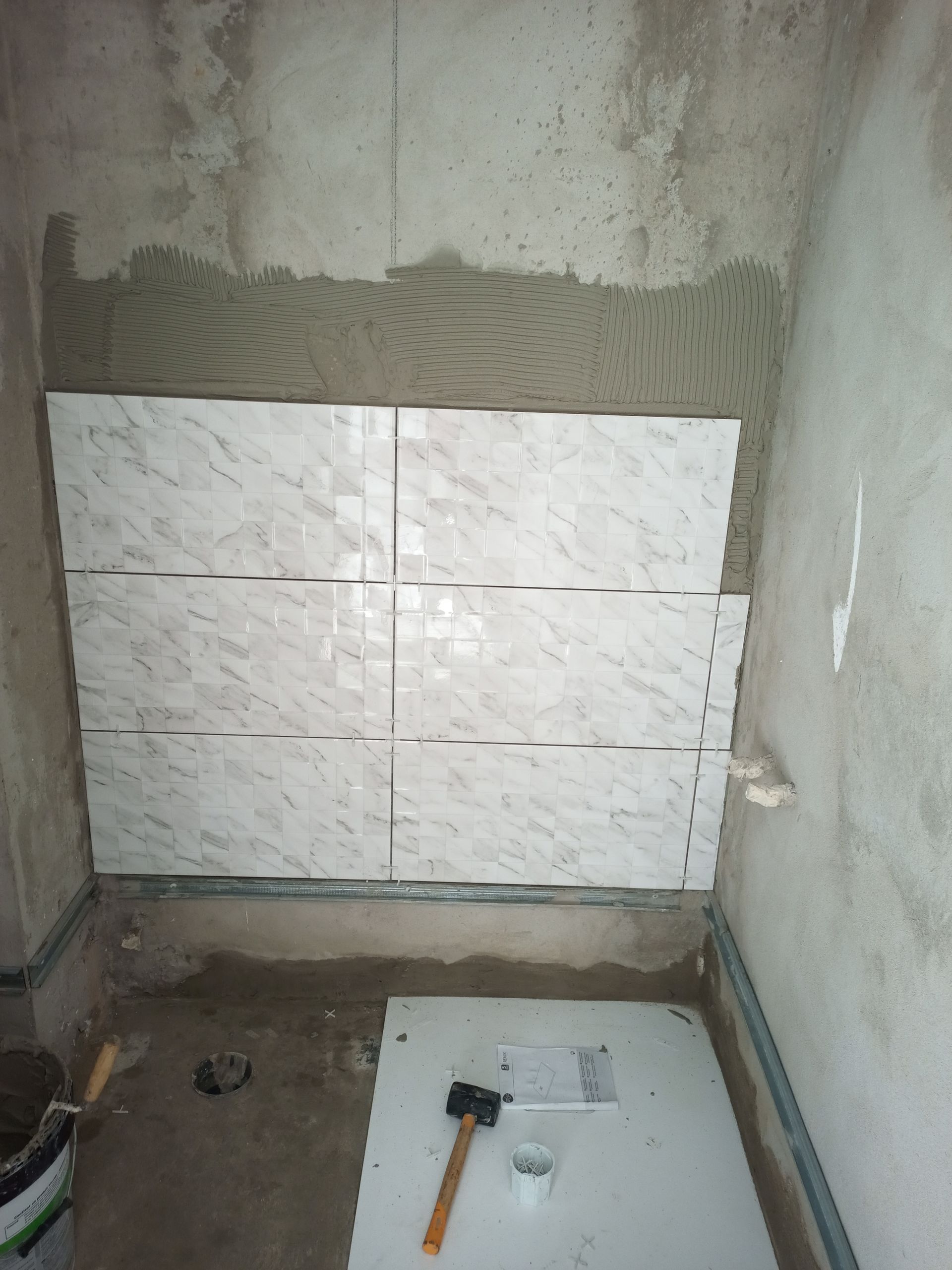 Colocação de azulejos em andamento; azulejos brancos sendo aplicados na parede com adesivo. Martelo e ferramentas visíveis nas proximidades.