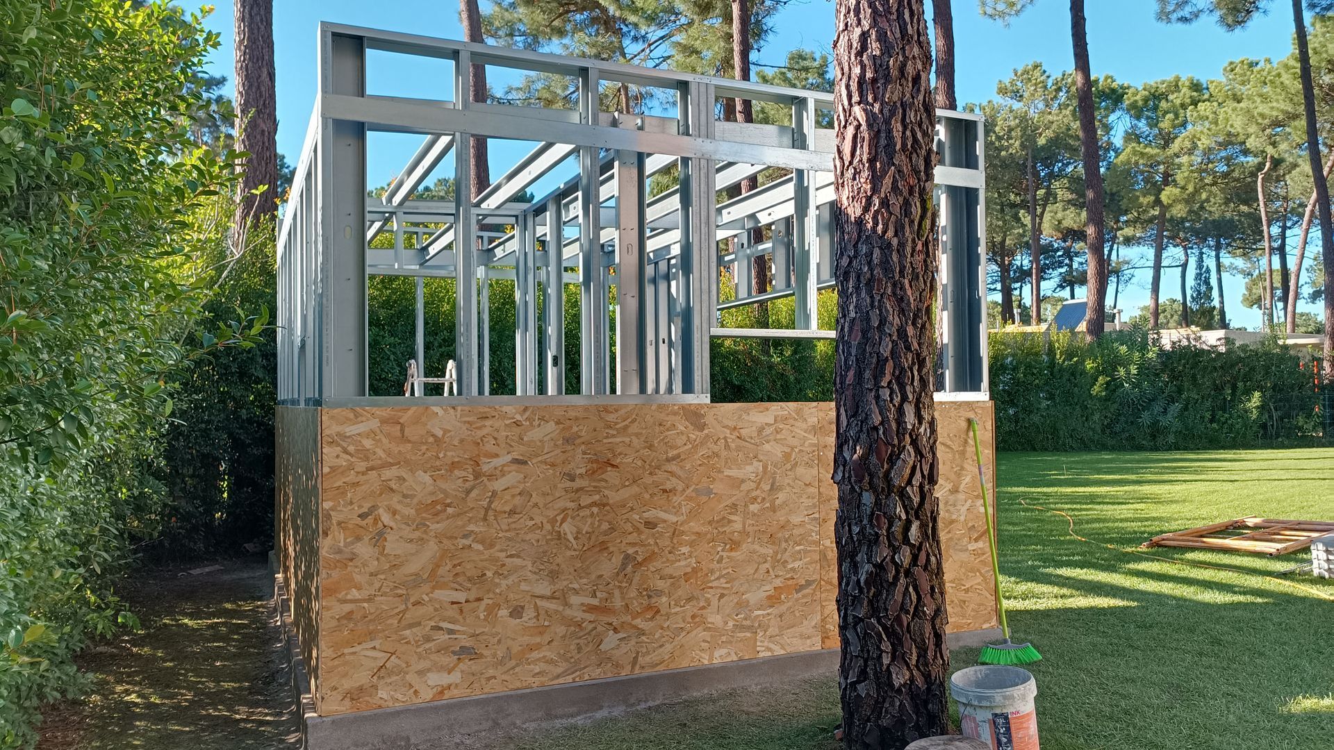 Estrutura parcialmente construída com painéis de madeira e vigas metálicas, em uma área gramada com árvores.