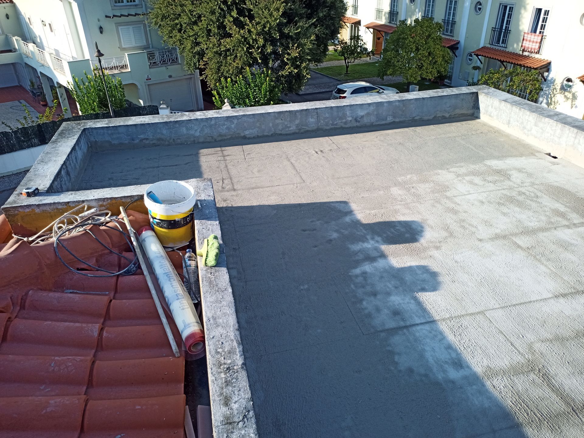 Telhado plano de concreto sendo trabalhado com ferramentas e um balde; telhas de terracota também são visíveis.