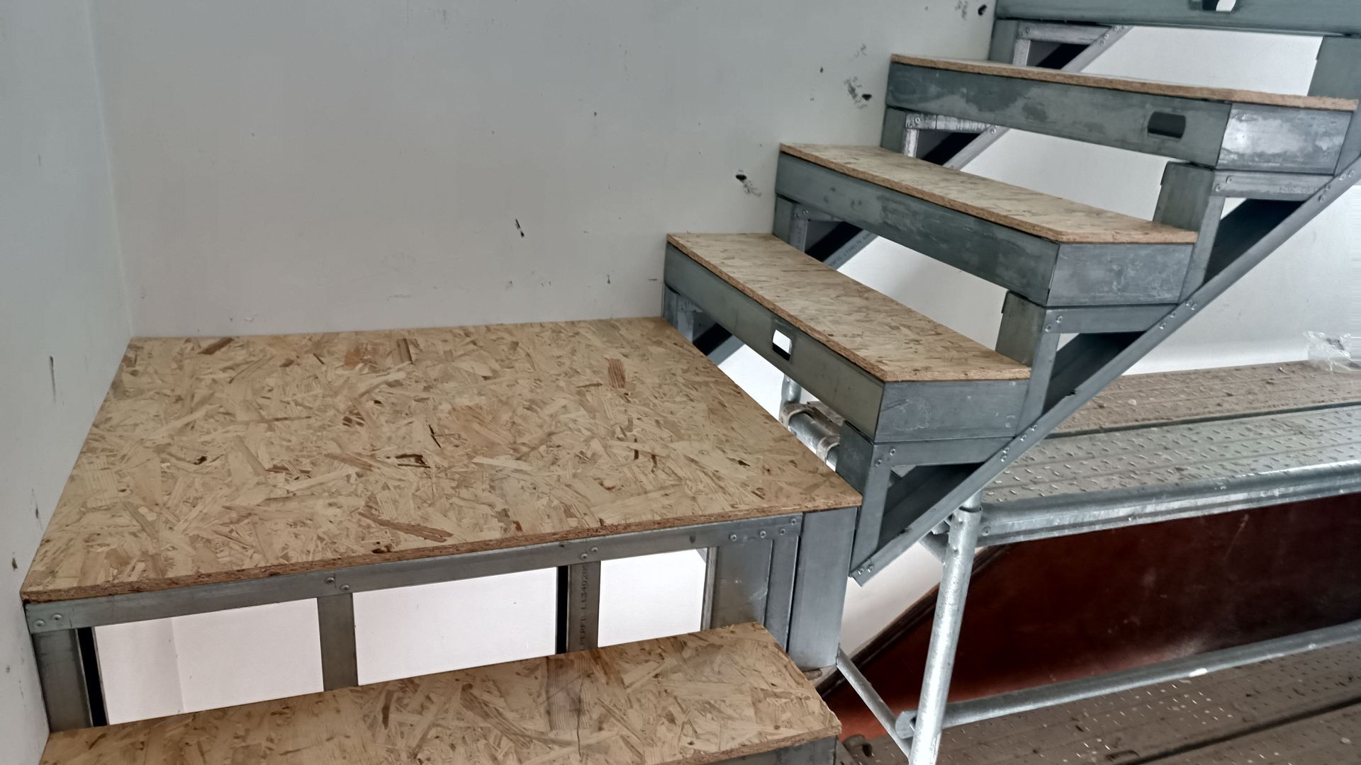 Escada com estrutura metálica e degraus de madeira compensada em construção contra uma parede branca.