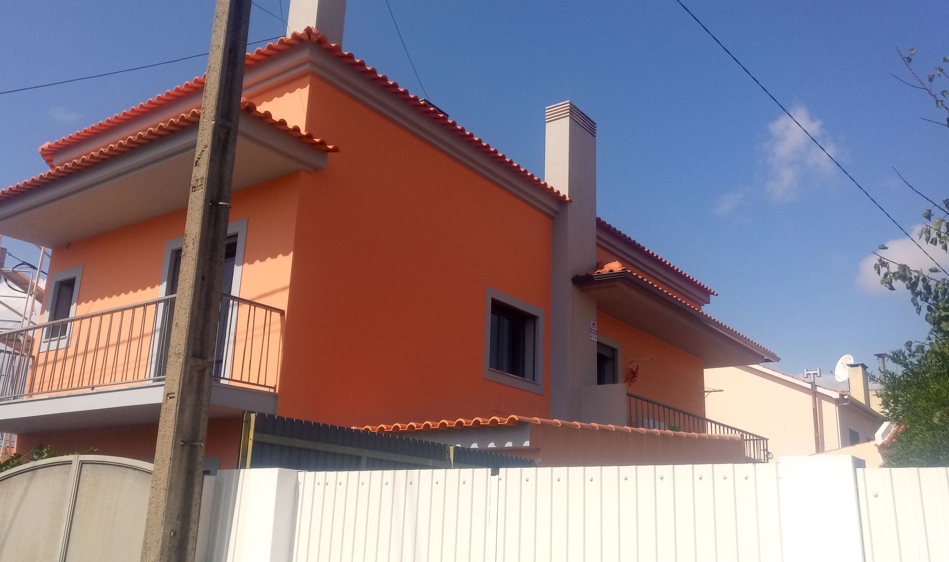 Prédio laranja com telhado vermelho, duas chaminés e varanda branca contra um céu azul.