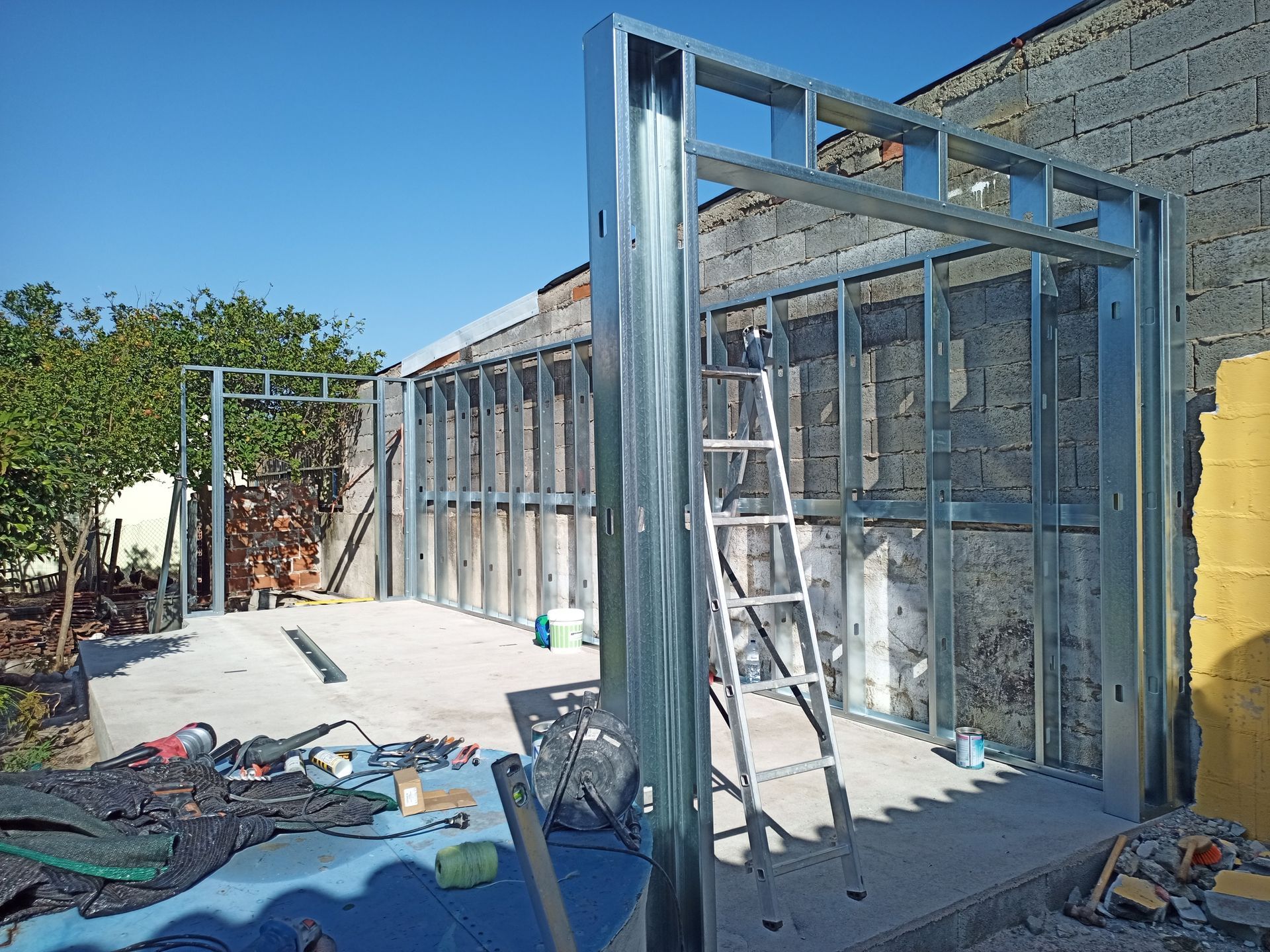 Estrutura metálica de um cômodo em construção encostada em uma parede de tijolos, com escada e ferramentas presentes.