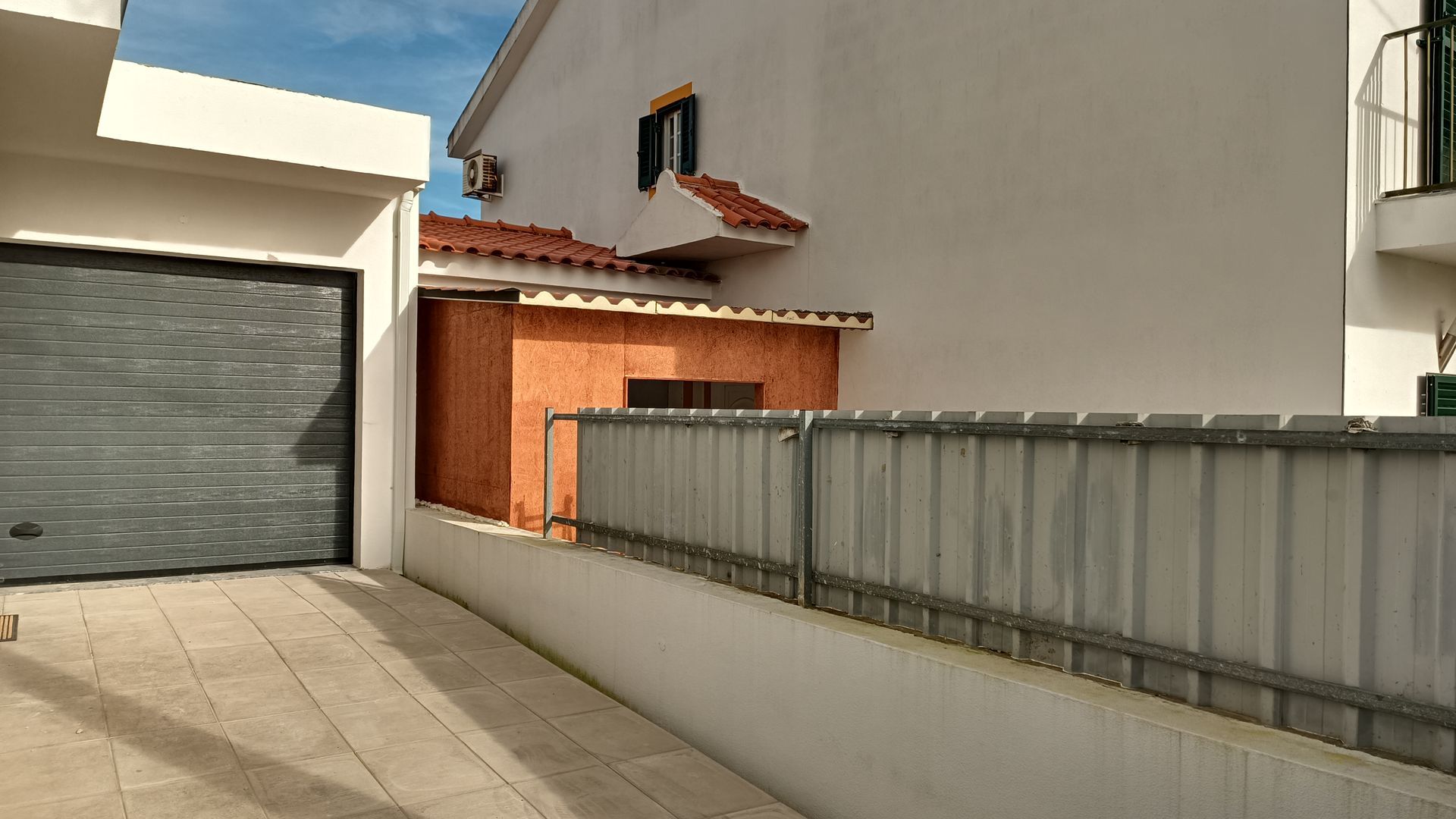 Porta de garagem cinza ao lado de um prédio branco e um galpão laranja; céu claro.