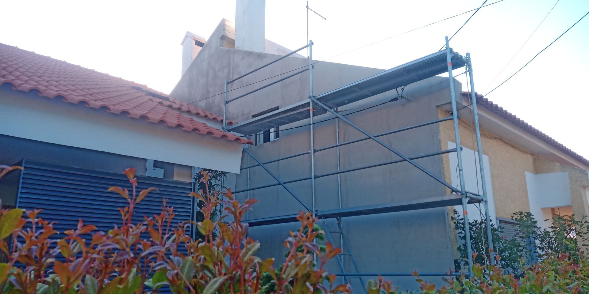 Andaimes ao lado de um prédio, possivelmente em reforma. Telhado vermelho, chaminé e plantas em primeiro plano.