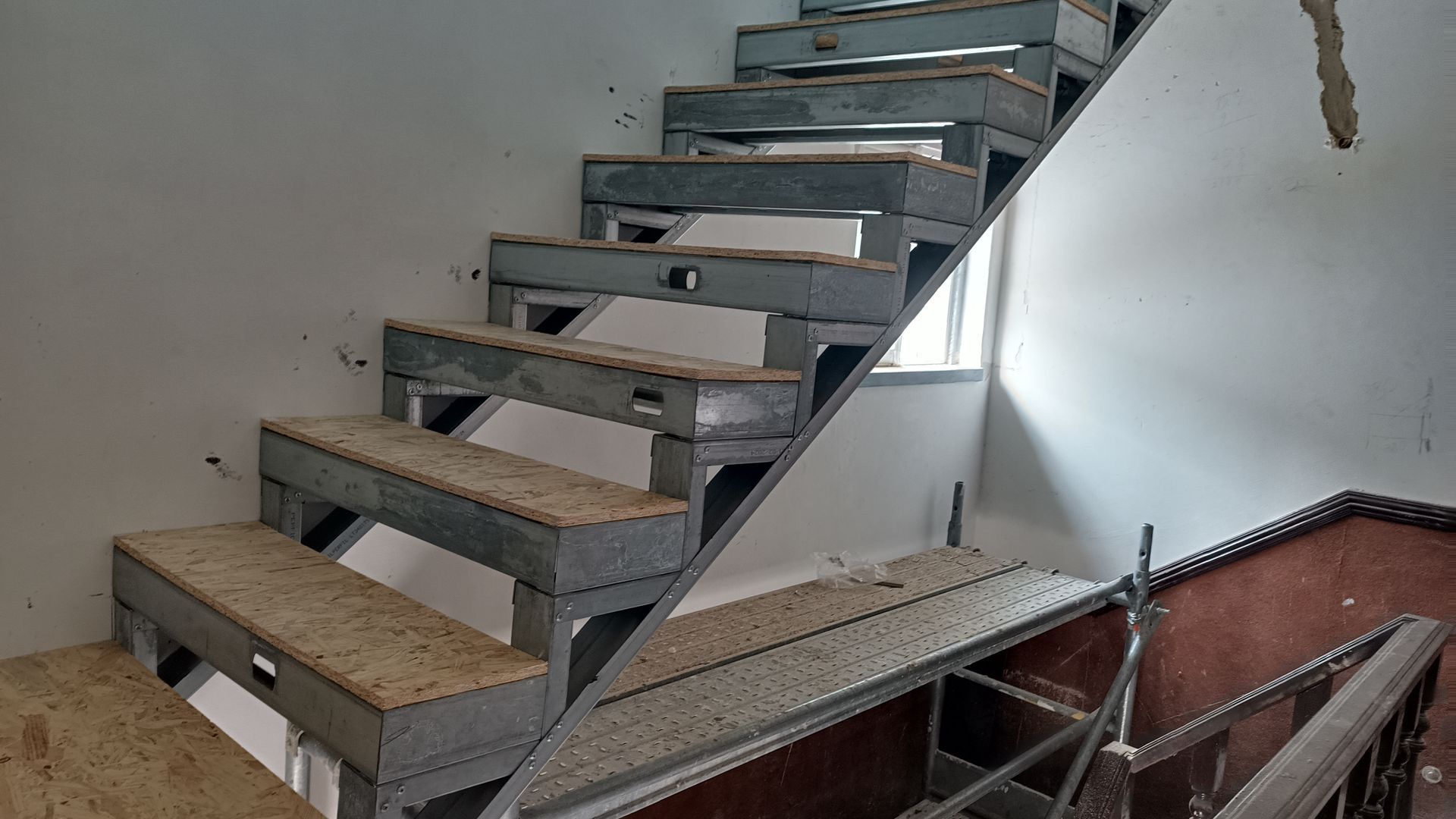 Escada metálica com degraus vazados subindo ao longo de uma parede branca, com uma janela ao fundo.