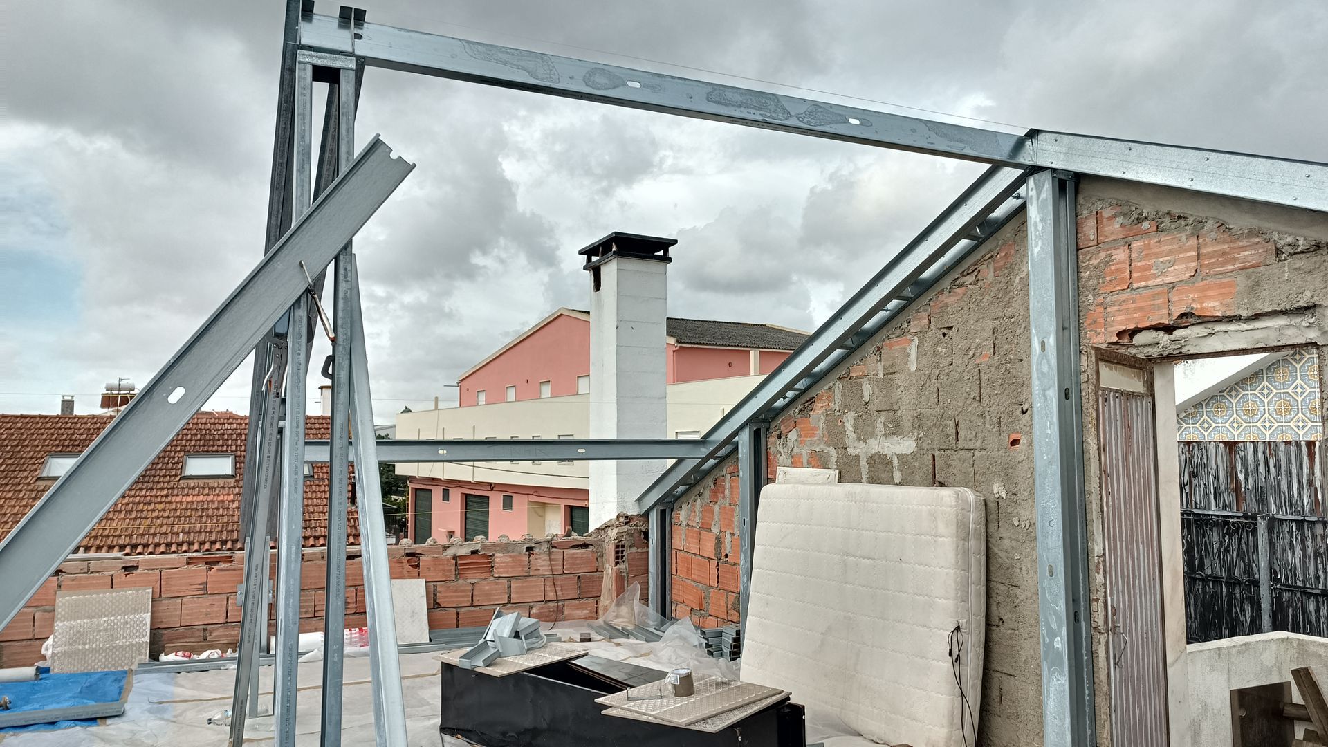 Estrutura metálica sobre um telhado, tijolos, chaminé, céu nublado.