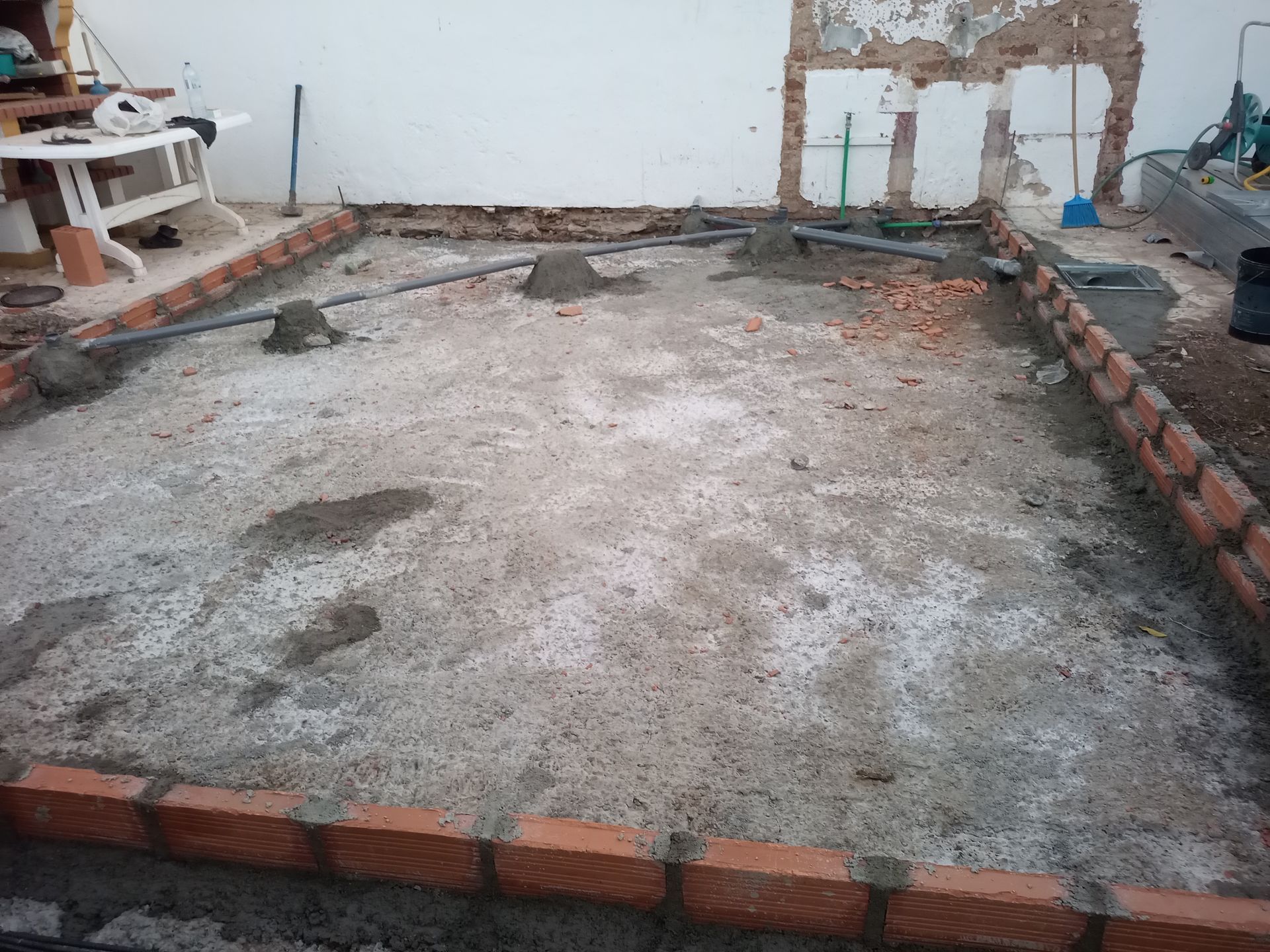 Canteiro de obras: Borda de tijolos ao redor de uma fundação de concreto, diversos materiais de construção e uma parede desgastada ao fundo.