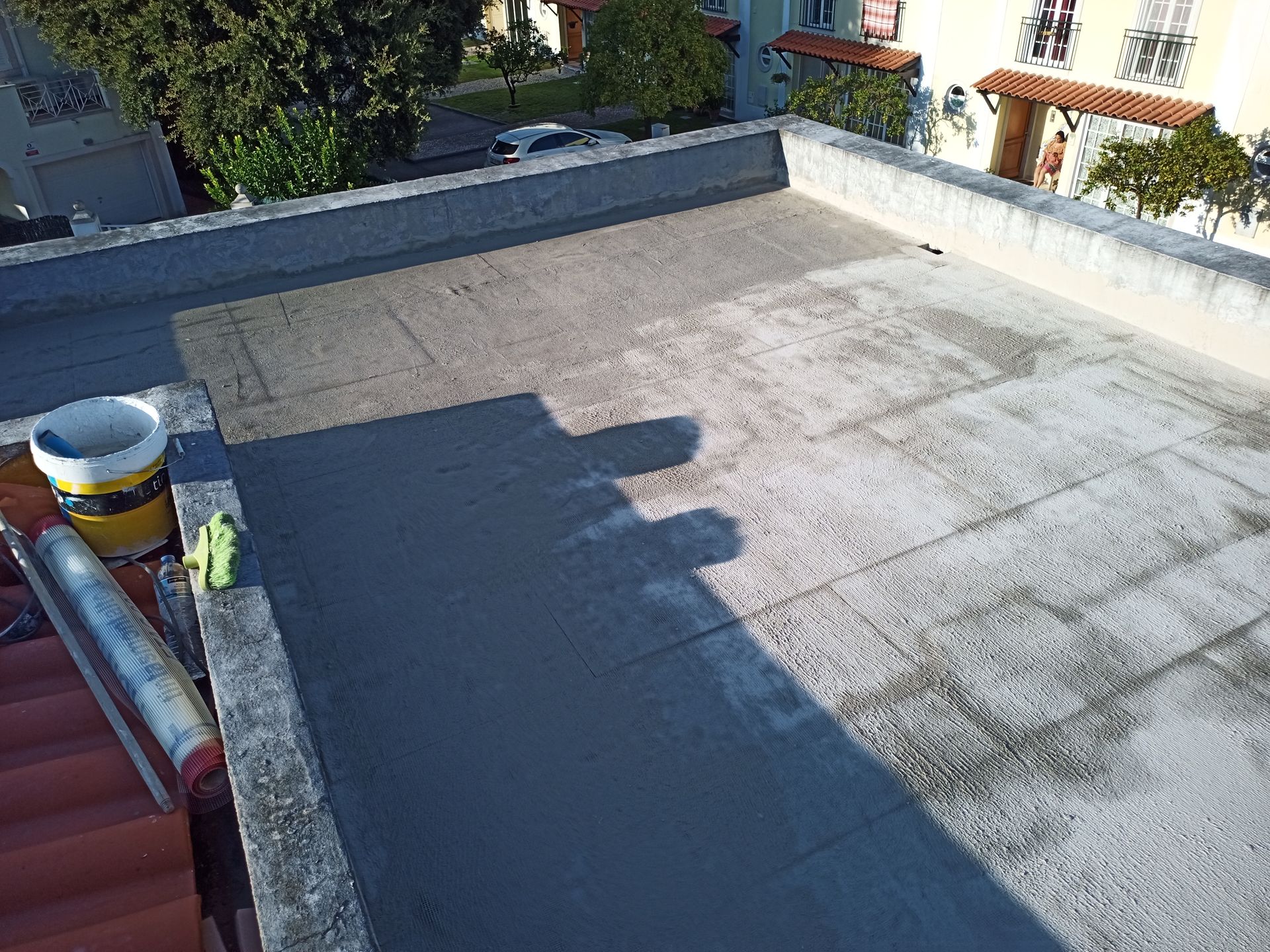 Telhado plano parcialmente coberto com selante cinza escuro; ferramentas e balde na borda.