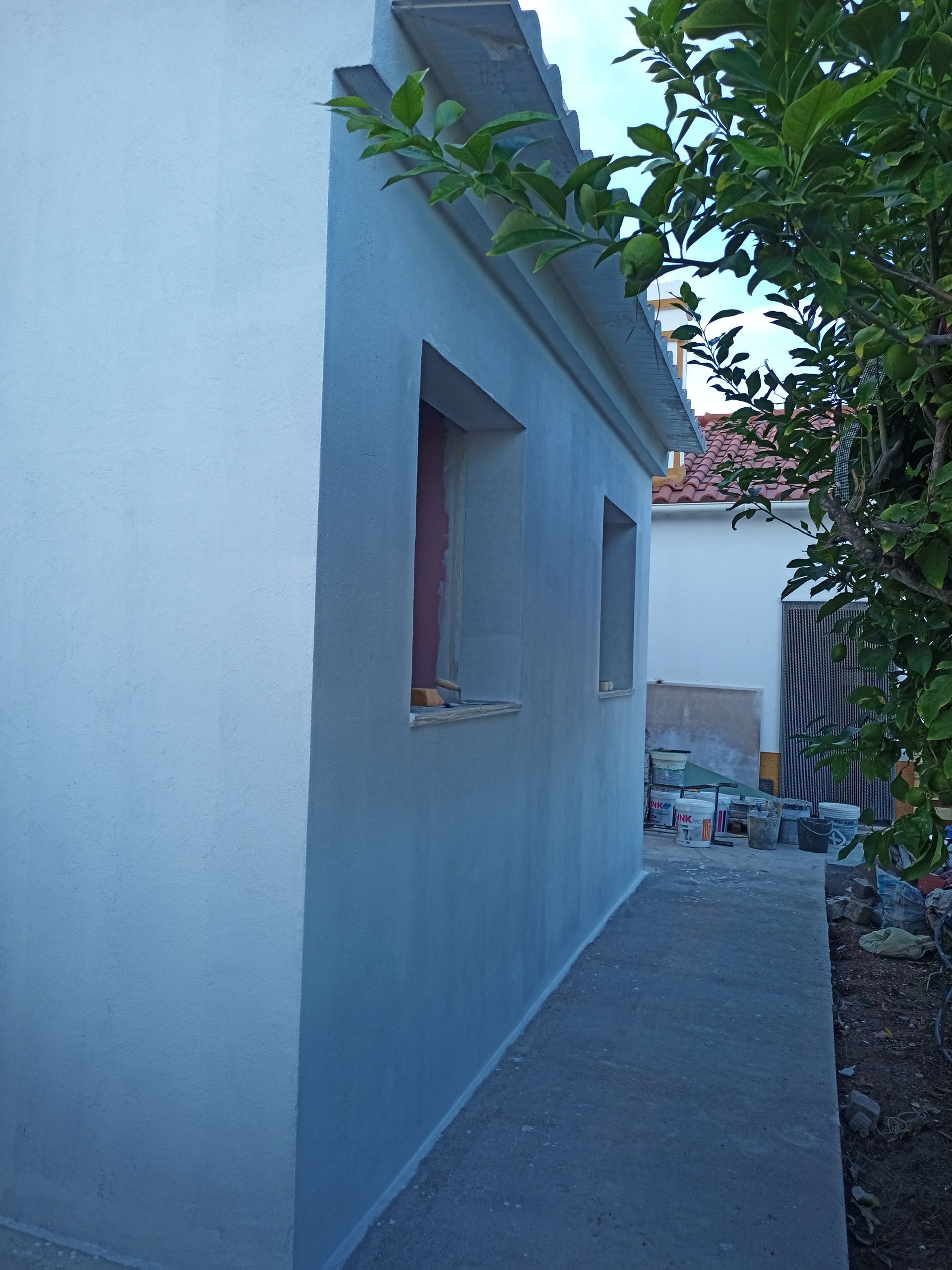 Fachada branca do prédio com duas janelas, uma passarela de concreto e vegetação.