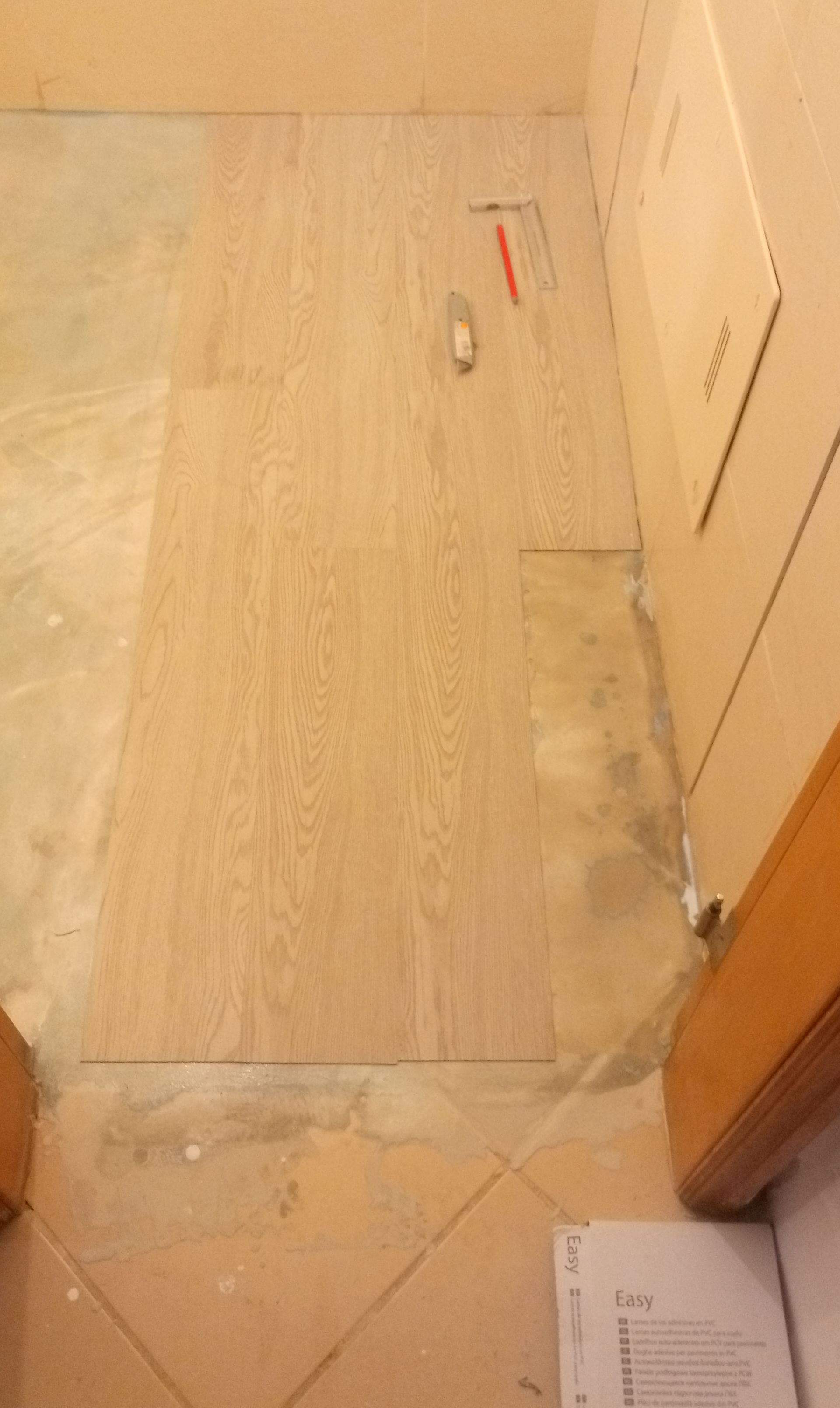 Piso de madeira sendo instalado próximo a um painel de acesso de utilidades e piso de cerâmica bege.