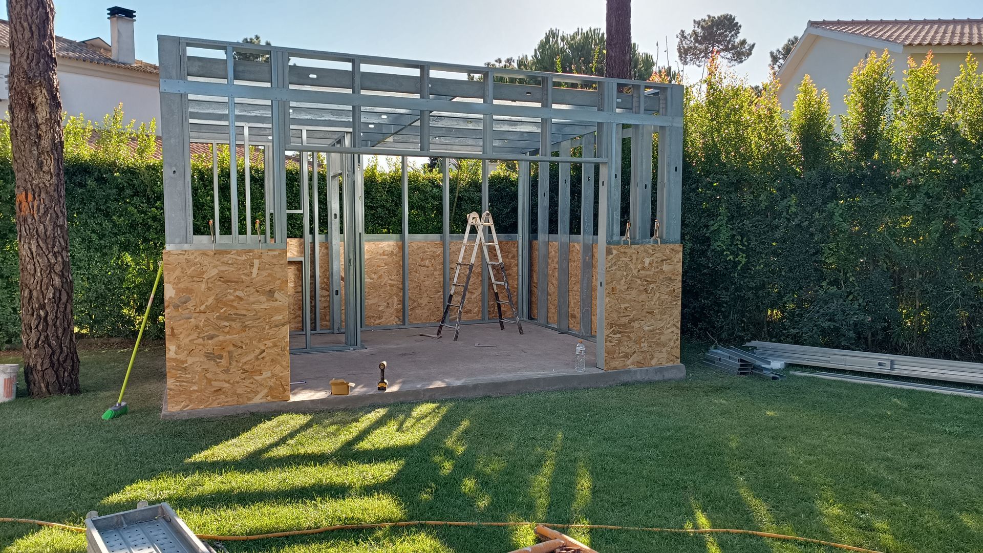 Estrutura metálica de uma construção externa parcialmente erguida, com base em aglomerado, sobre um gramado.