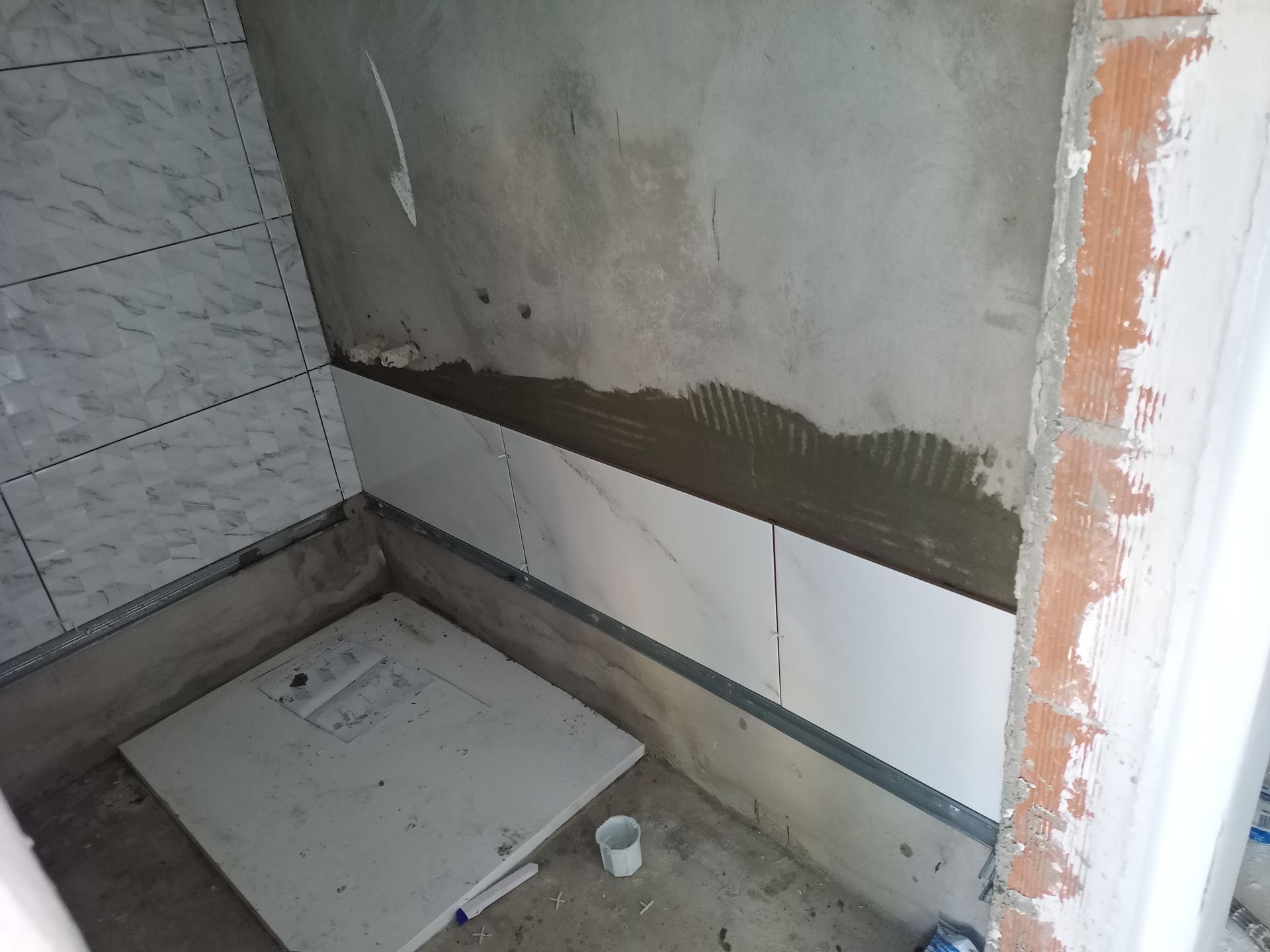 Construção de uma parede de chuveiro revestida com azulejos brancos com aparência de mármore. Cimento, metal e tijolo aparente também são visíveis.