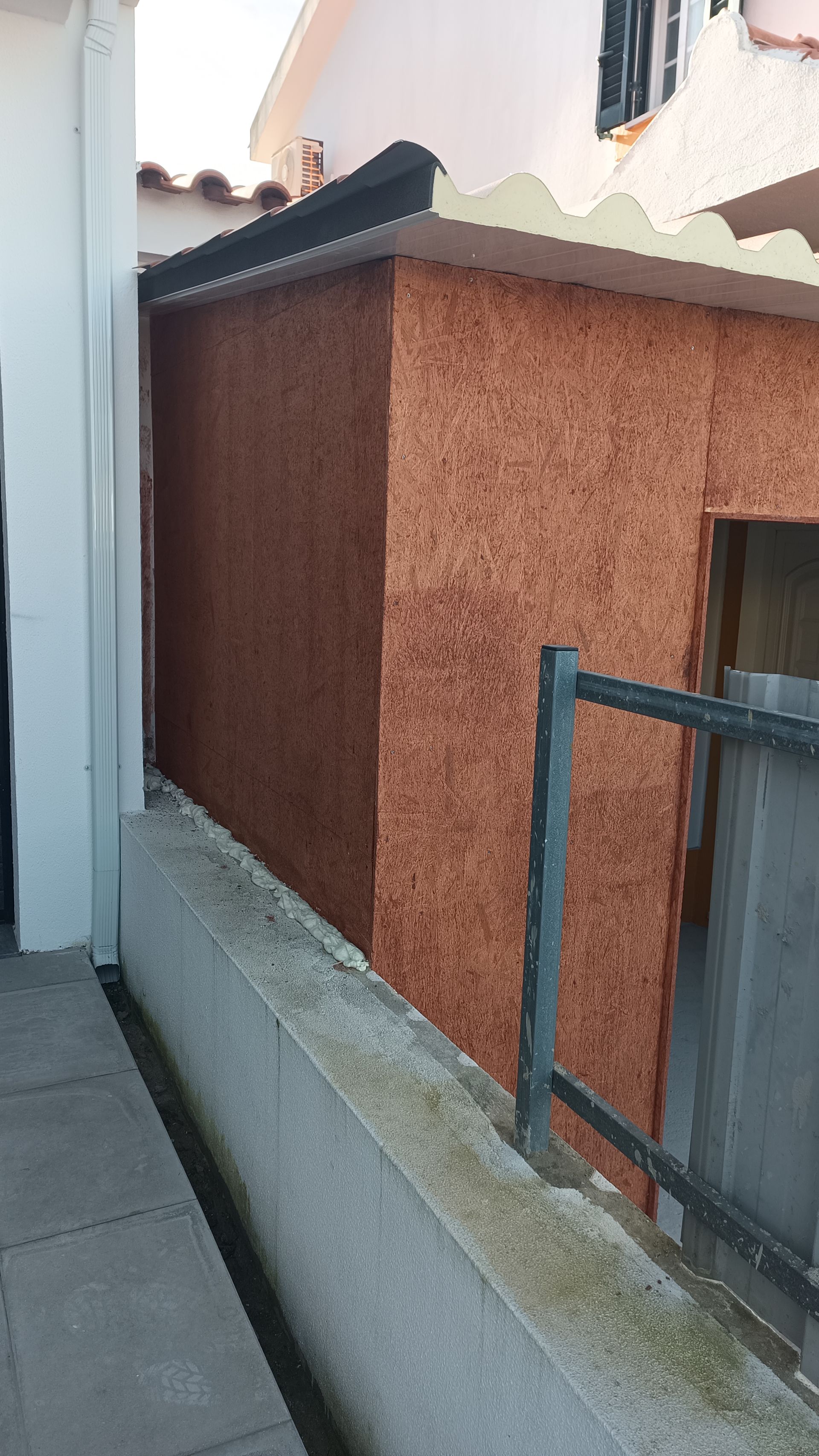 Um prédio estreito, com painéis marrons, telhado branco e cerca cinza.