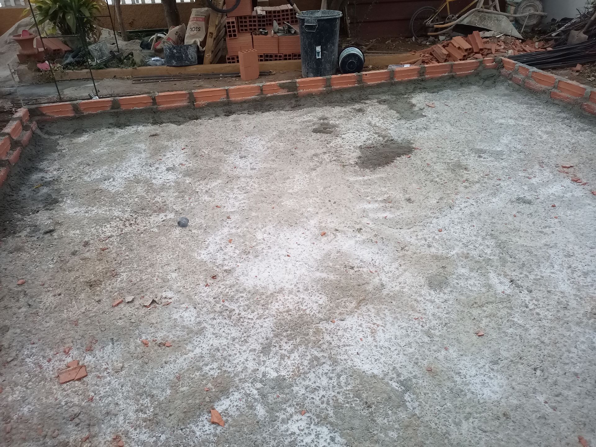 Placa de concreto com borda de tijolos em construção; tijolos e materiais espalhados ao fundo.