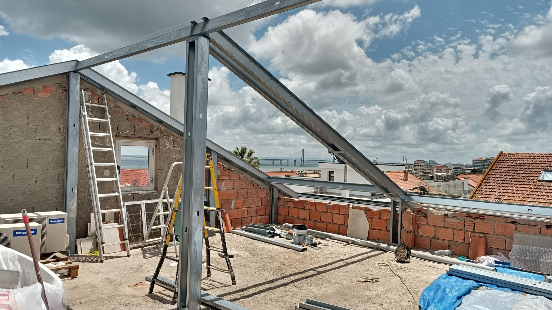 Canteiro de obras: estrutura metálica em um telhado, paredes de tijolos, escadas, céu nublado ao fundo.