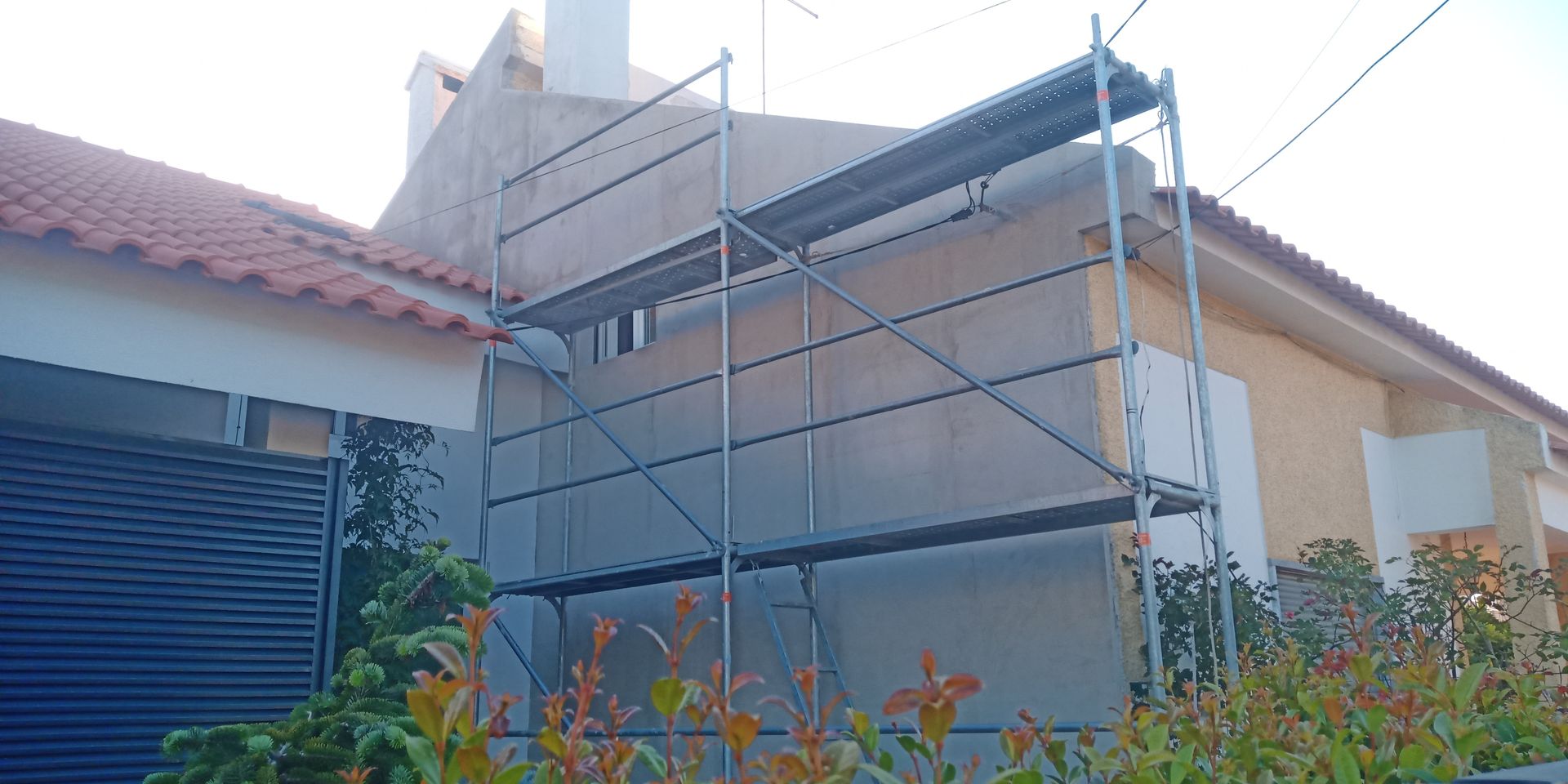 Andaimes encostados em um prédio em reforma; telhado de telhas laranja e arbustos embaixo.