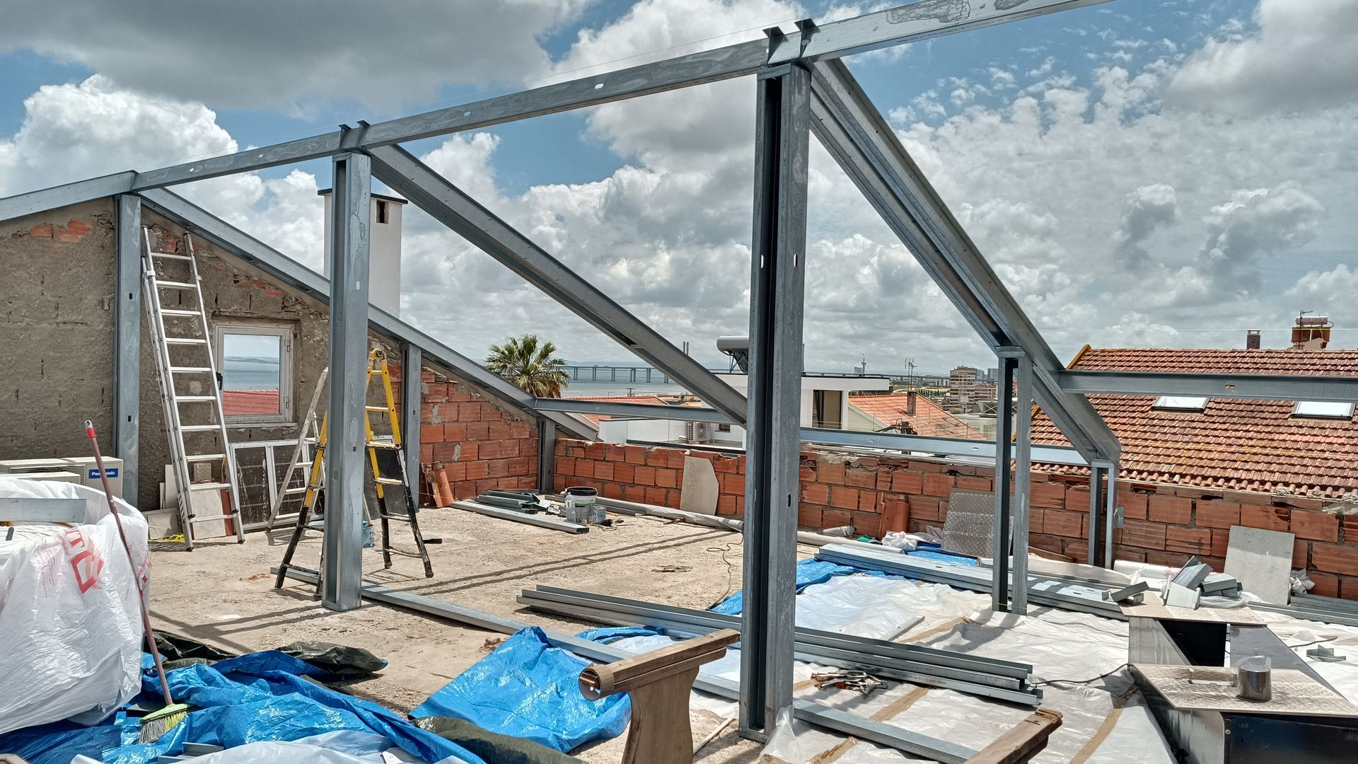 Canteiro de obras com estrutura metálica em um telhado, paredes de tijolos, uma escada e céu nublado.