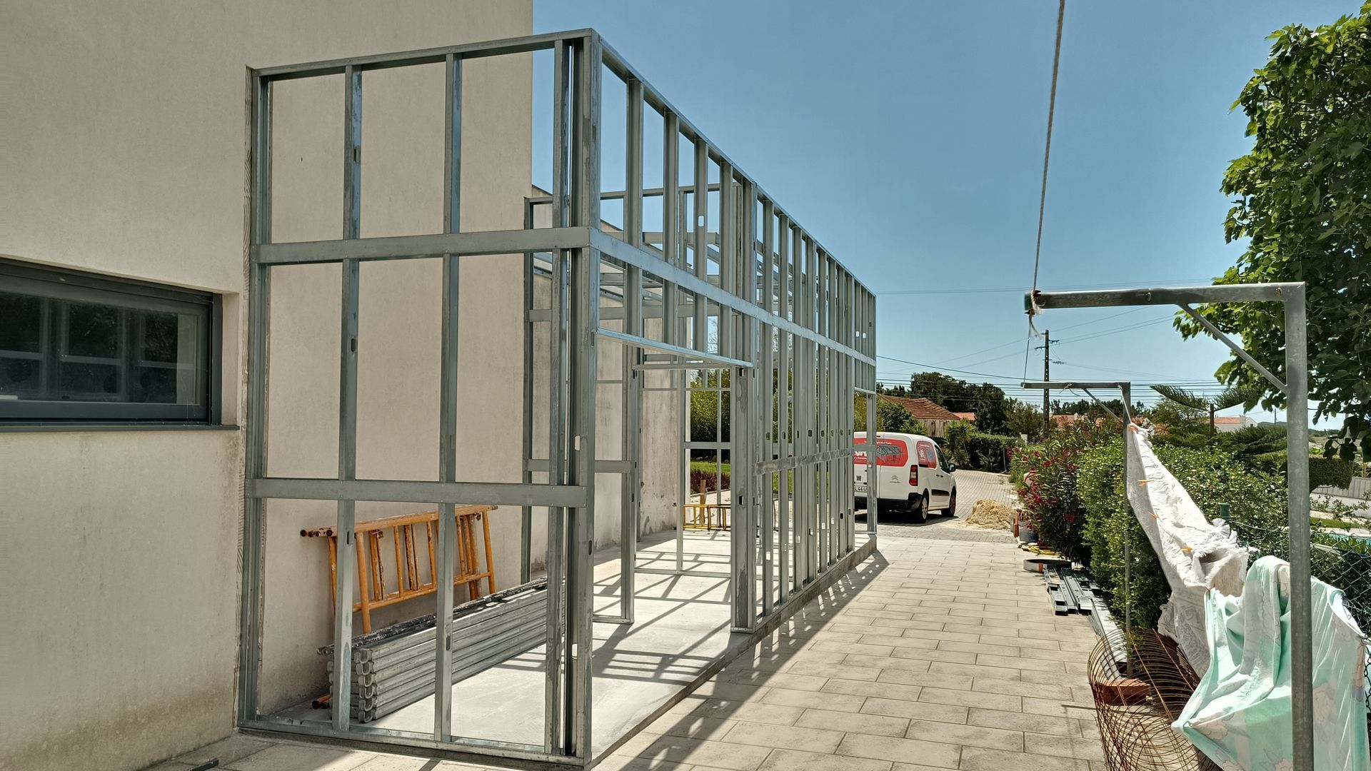 Estrutura metálica de um edifício em construção, fixada a uma parede branca com uma janela. Ambiente externo ensolarado.