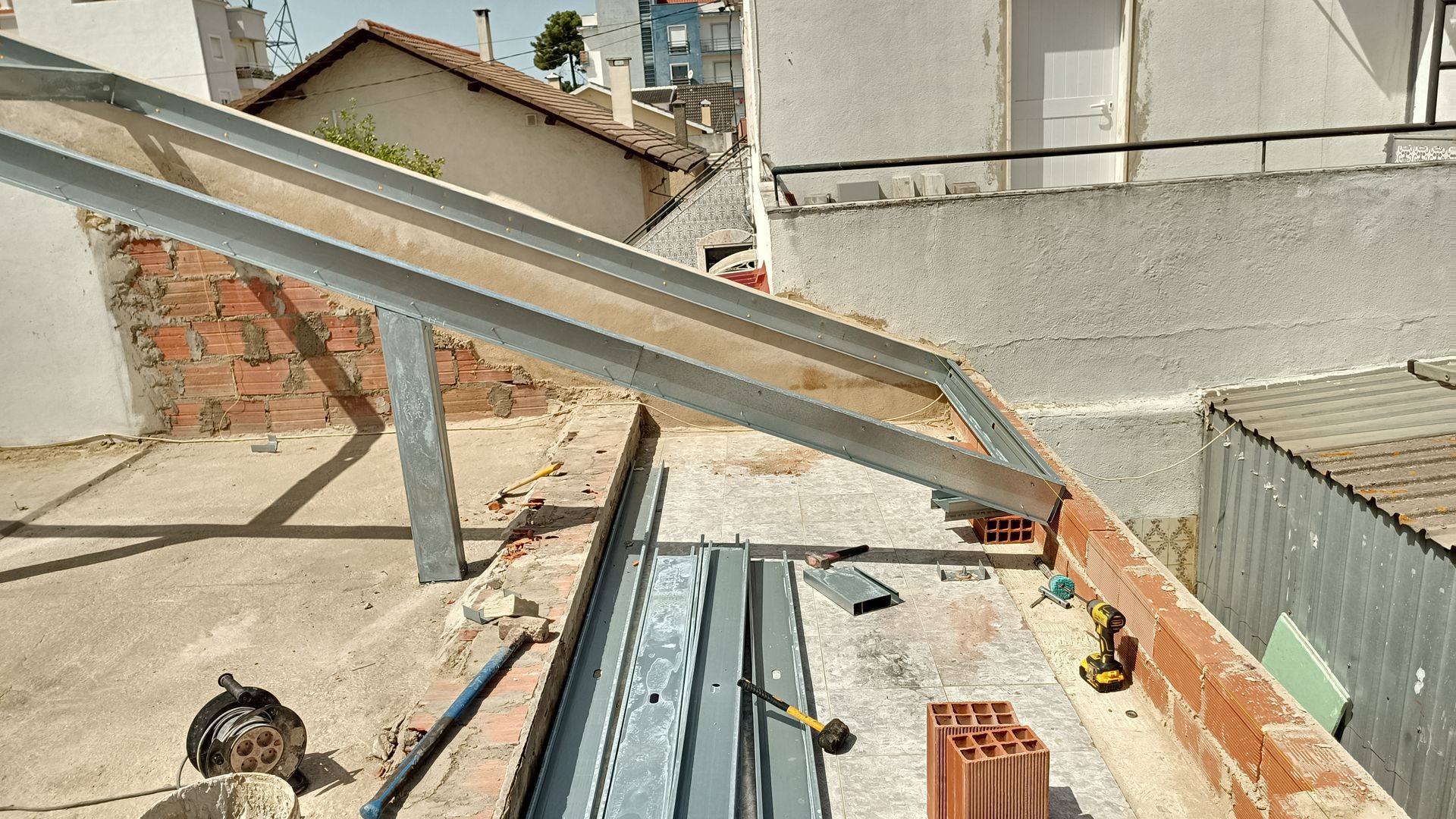 Cena de construção: estrutura metálica sobre parede de tijolos, ferramentas em um telhado, com prédios ao fundo.