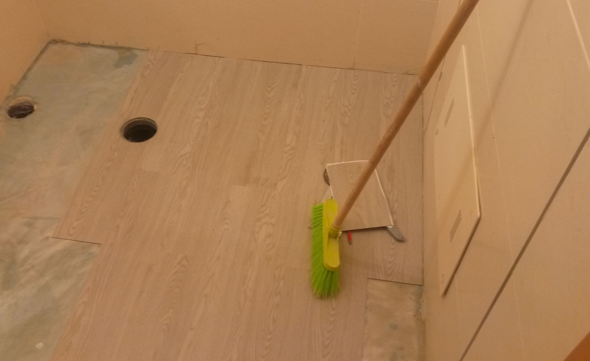 Uma pessoa varre um piso de azulejos com uma escova verde e amarela, em uma sala de cores neutras.