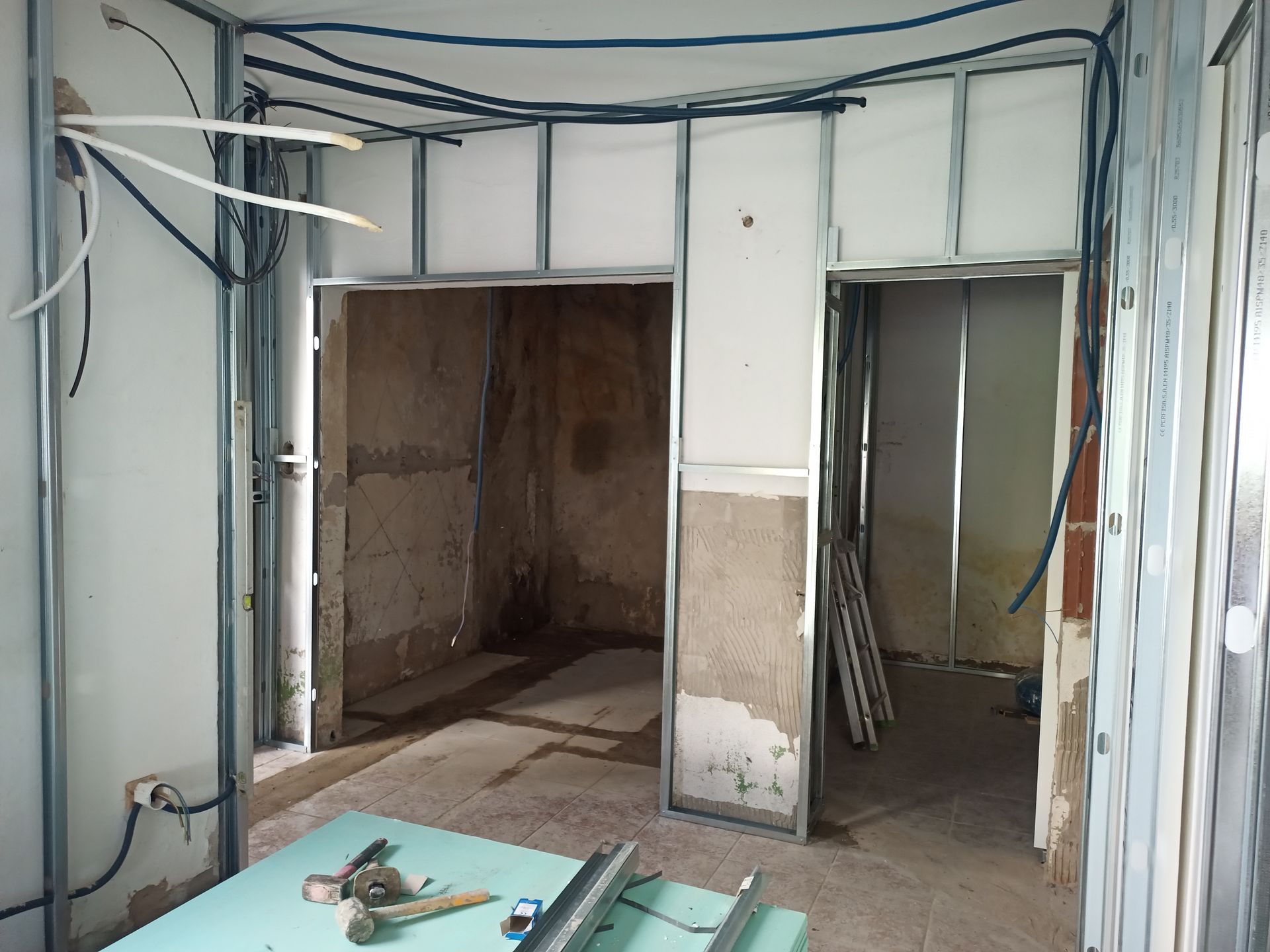 Quarto interno em construção, estrutura de metal, fiação exposta, duas portas, entulho no chão, tons neutros.