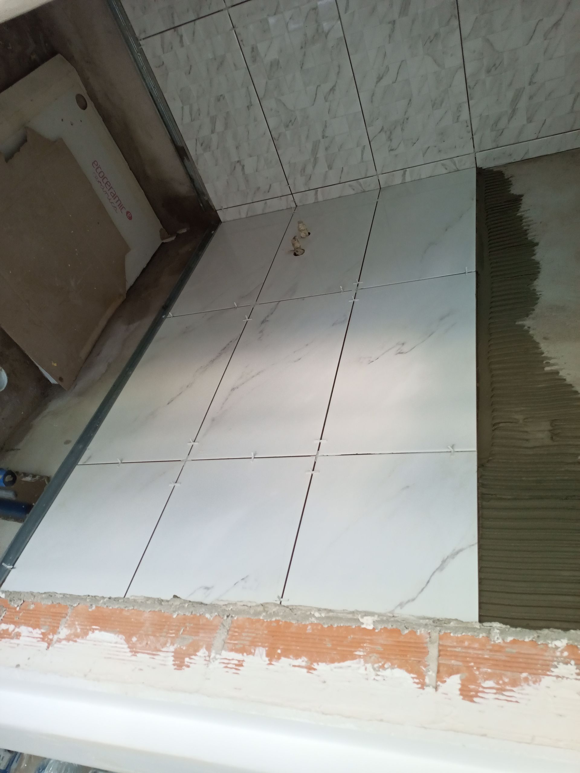 Piso e parede revestidos com azulejos brancos em construção; mostrando argamassa, espaçadores e bordas inacabadas.