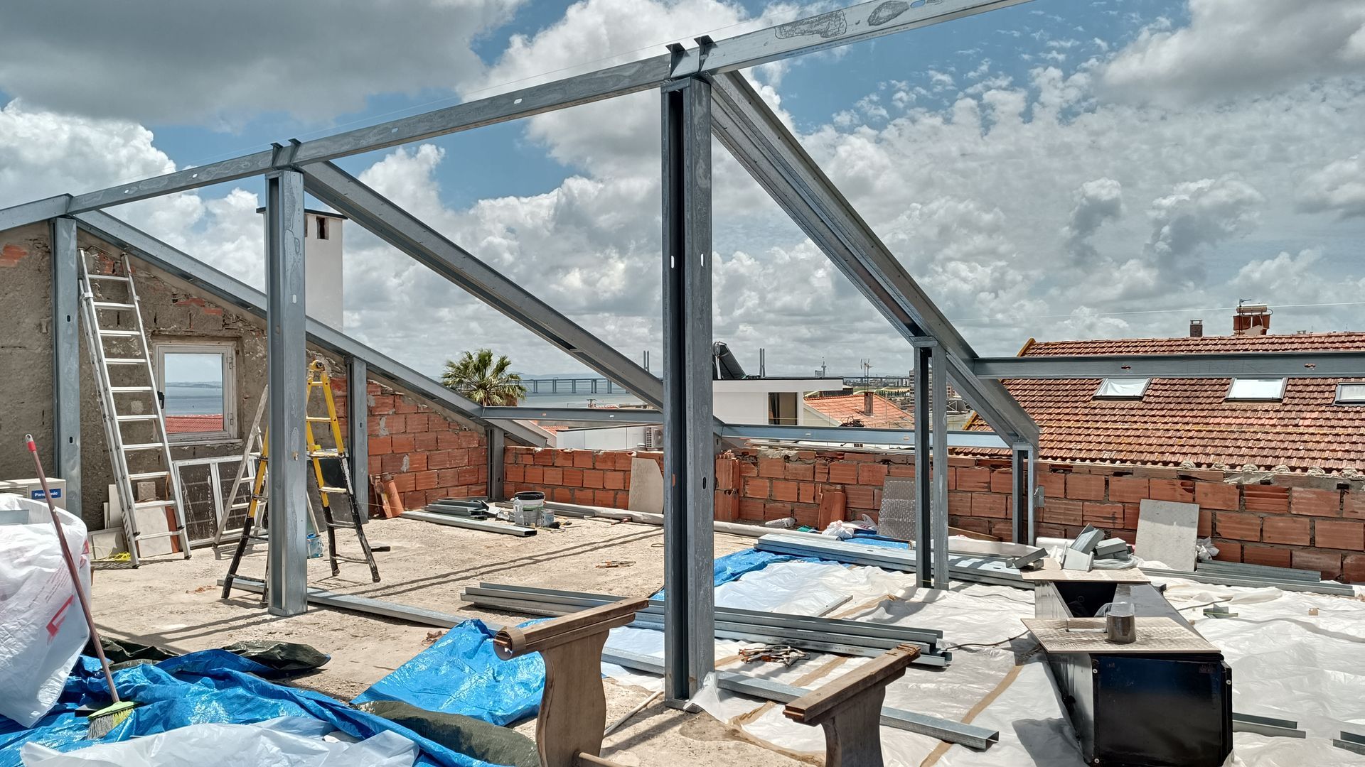 Vigas de aço formam a estrutura do telhado em uma cobertura em construção. Tijolos e ferramentas são visíveis.