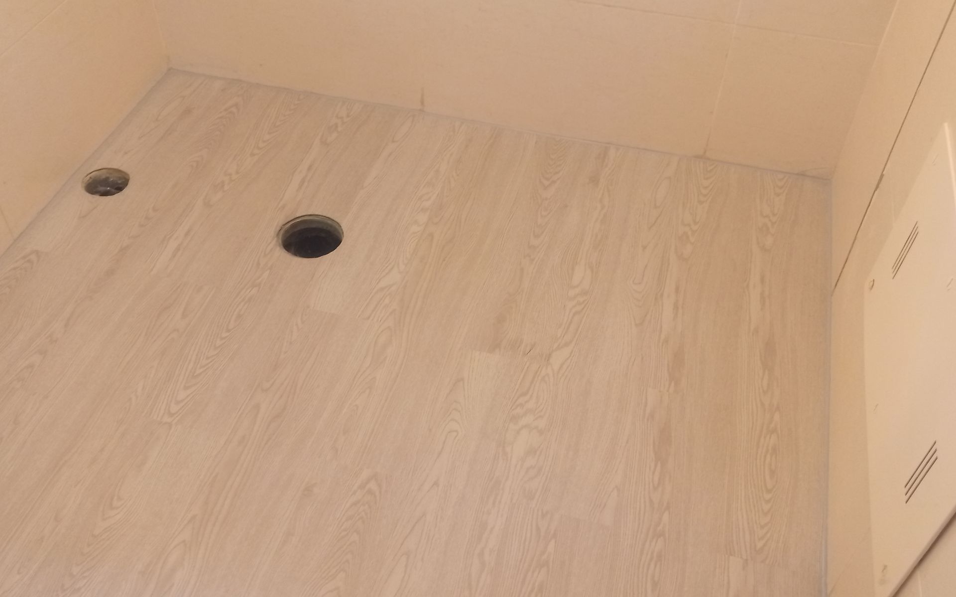 Quarto vazio com piso de madeira clara e dois buracos no chão, paredes bege.