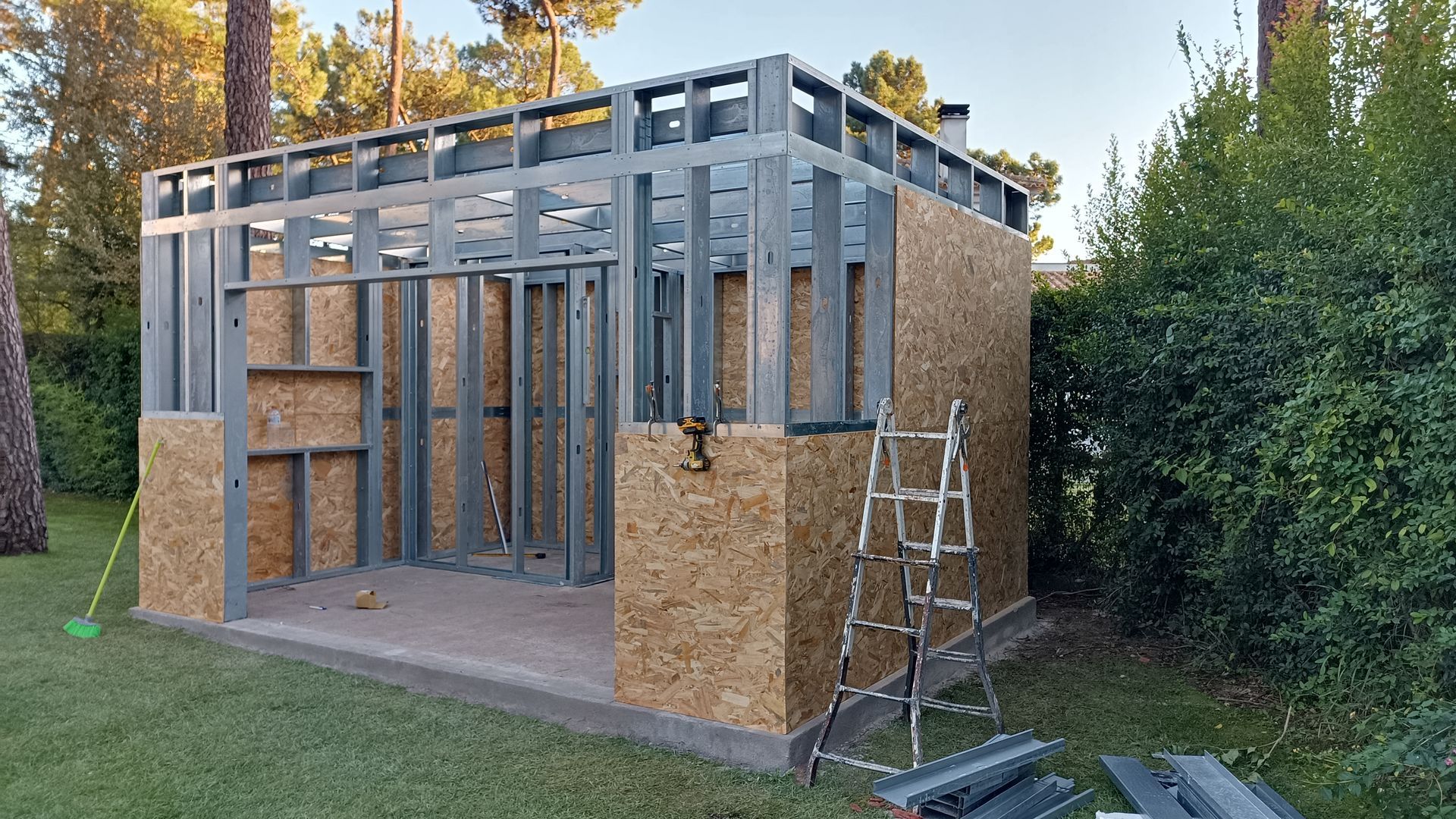 Edifício com estrutura de aço em construção, parcialmente revestido com painéis de madeira. Uma escada está posicionada nas proximidades, em um gramado.