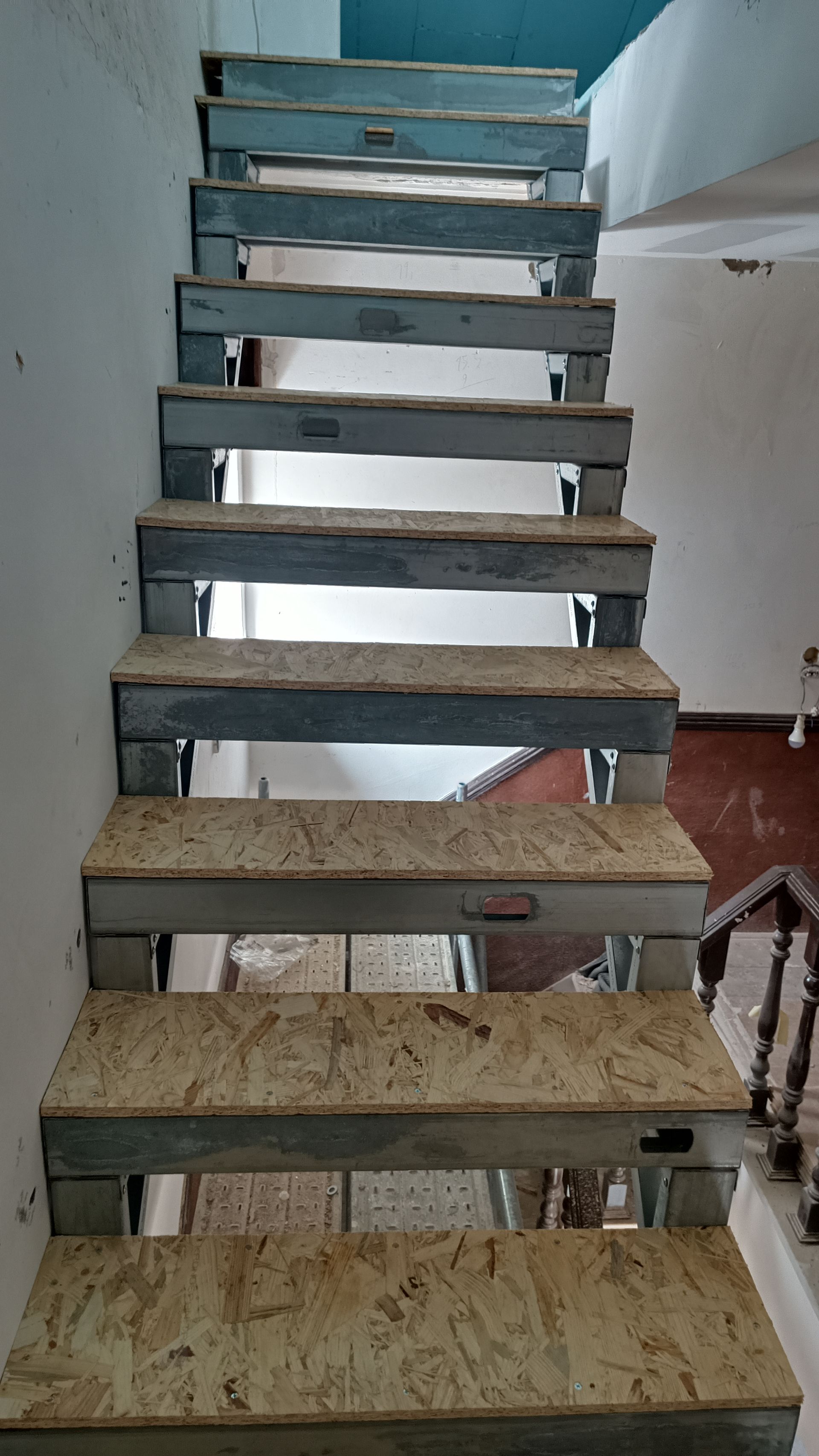 Escada com degraus de madeira e suportes de metal, subindo verticalmente contra uma parede de cor clara.