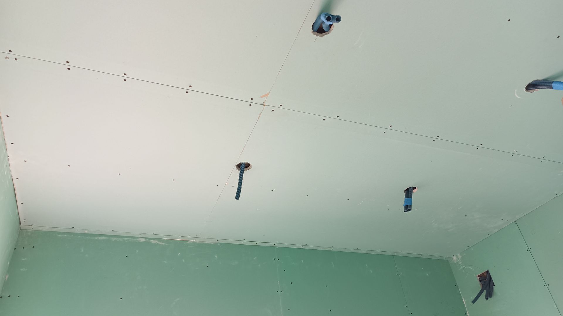 Teto de gesso cartonado com encanamento aparente e furos de parafusos, em uma sala com paredes verdes.