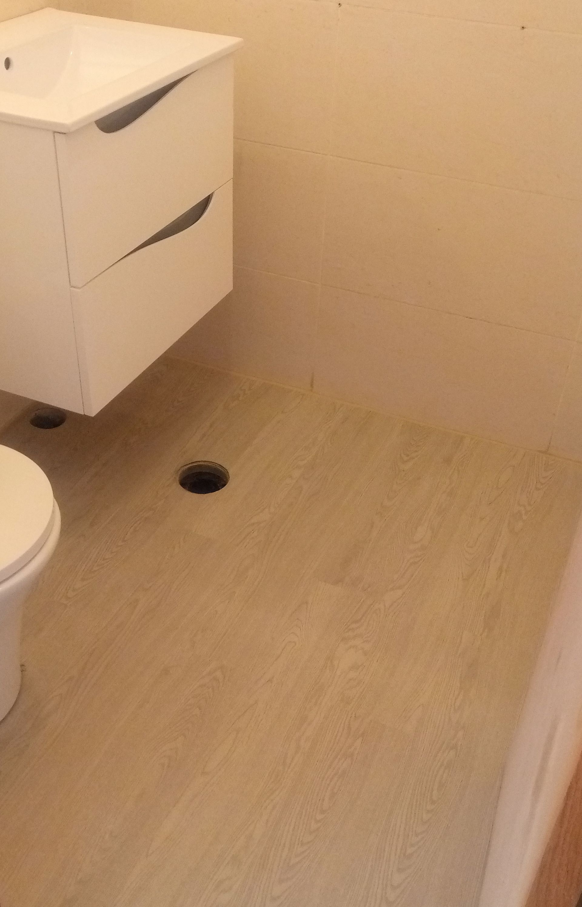 Banheiro com pia e vaso sanitário brancos, piso bege e ralos abertos.