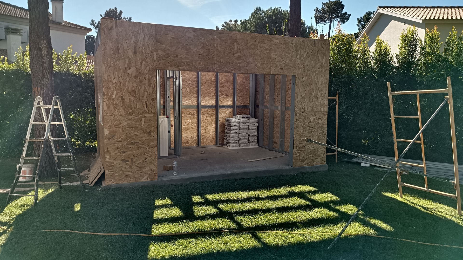 Galpão em construção em um quintal gramado; paredes de OSB, estrutura metálica, andaimes e escada.