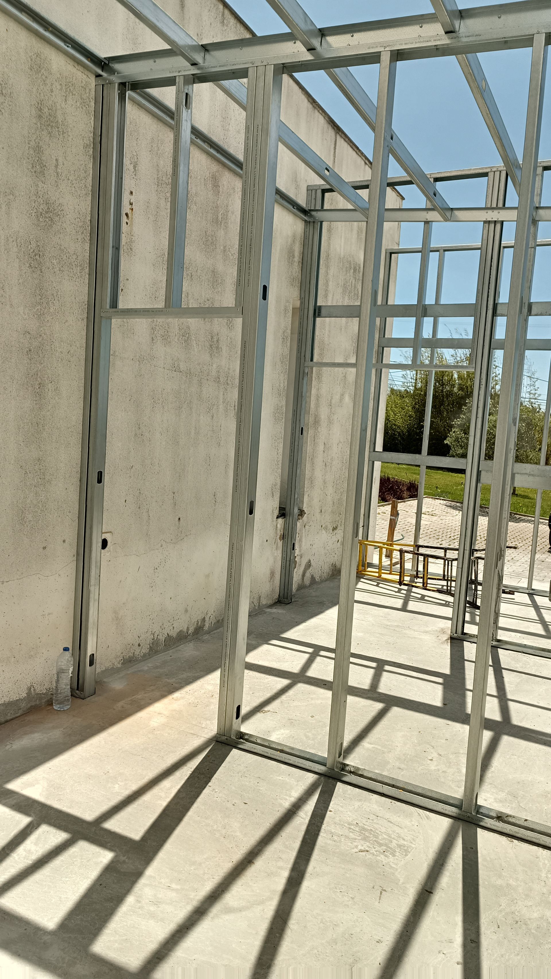 Estrutura metálica para paredes internas; a luz do sol projeta sombras no piso de concreto.