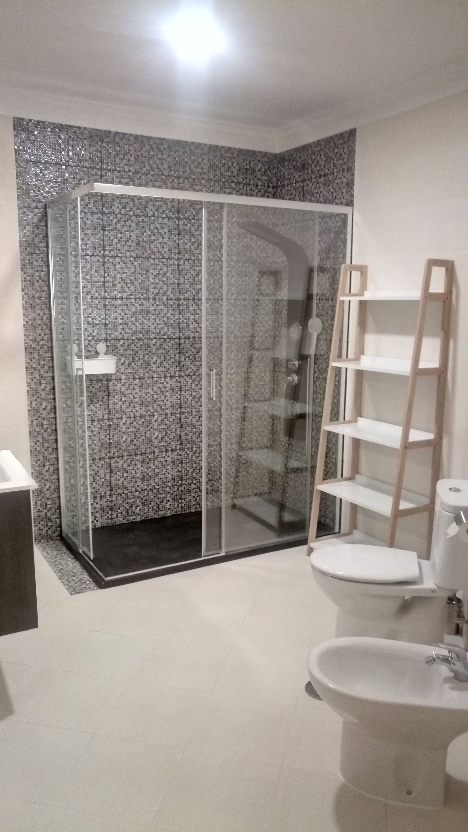 Banheiro com box de vidro, parede revestida com azulejos em mosaico, vaso sanitário e bidê brancos, e prateleira.