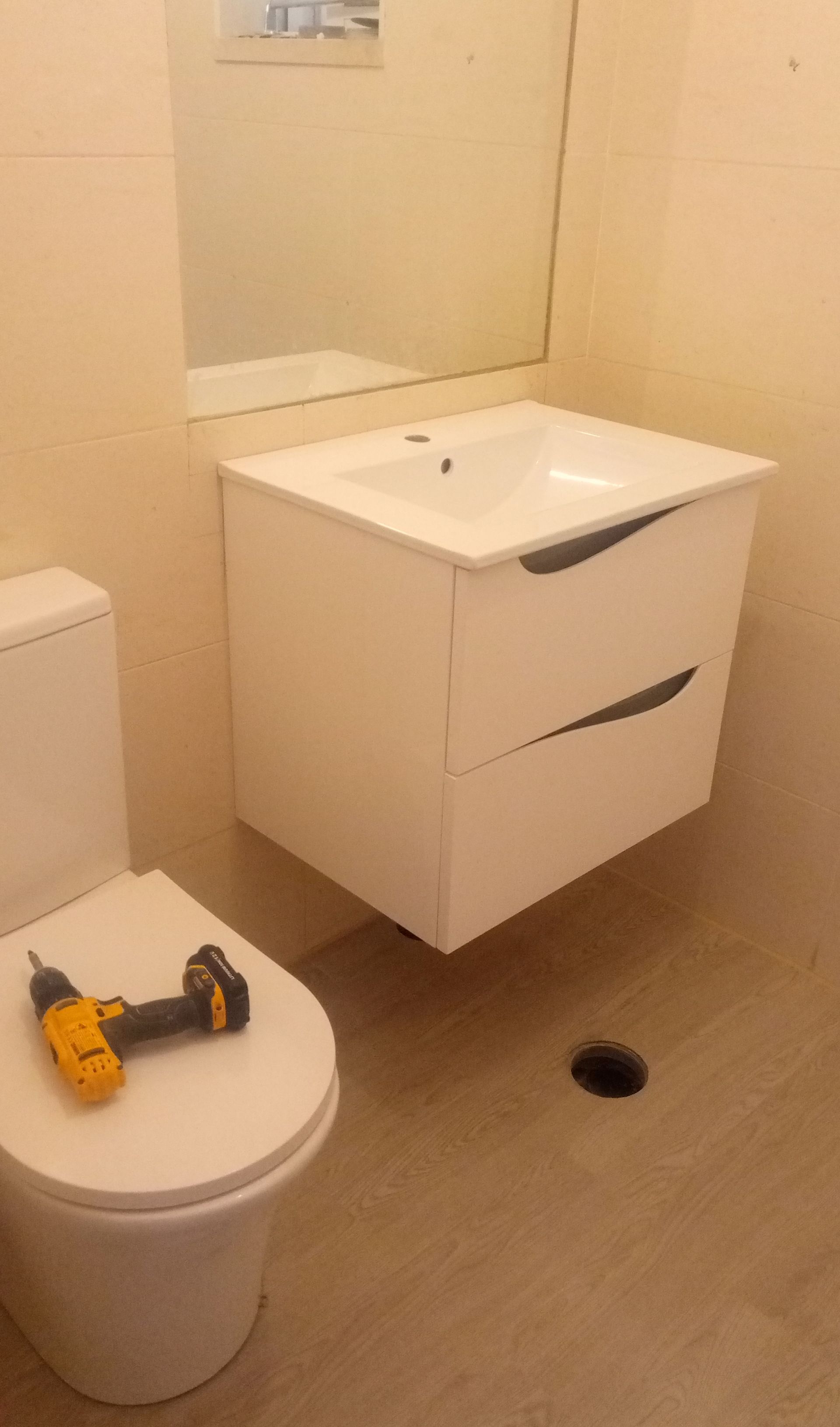 Móvel de banheiro suspenso branco com duas gavetas e um vaso sanitário branco. Uma furadeira sem fio está apoiada no vaso sanitário.