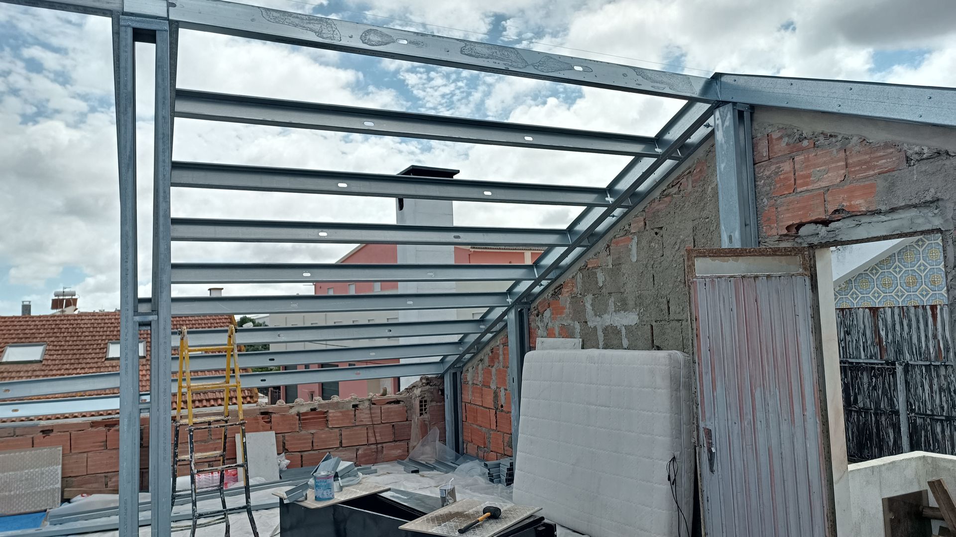 Estrutura metálica de um telhado parcialmente construído sobre um edifício de tijolos, sob um céu nublado.