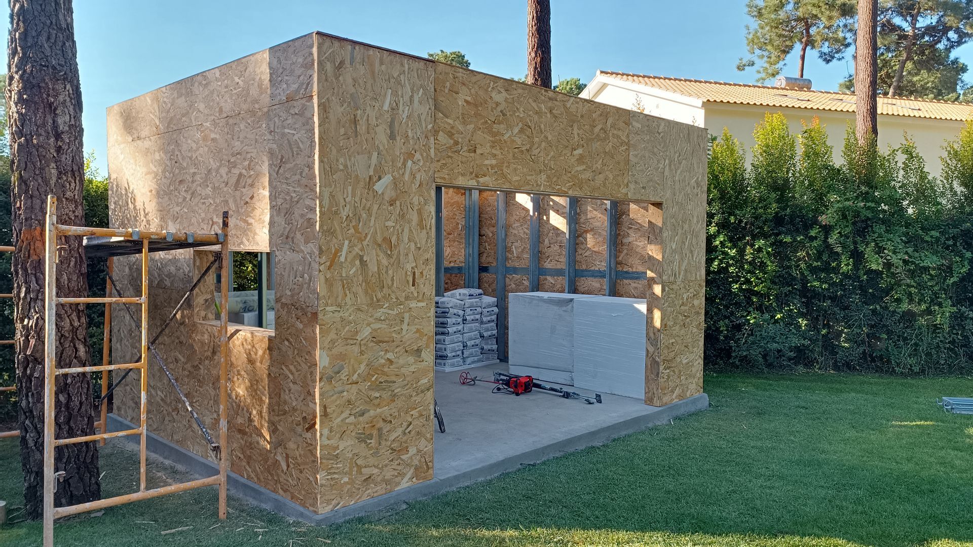 Construção de um pequeno galpão retangular com paredes de OSB, uma abertura para porta e interior parcialmente acabado em um gramado.